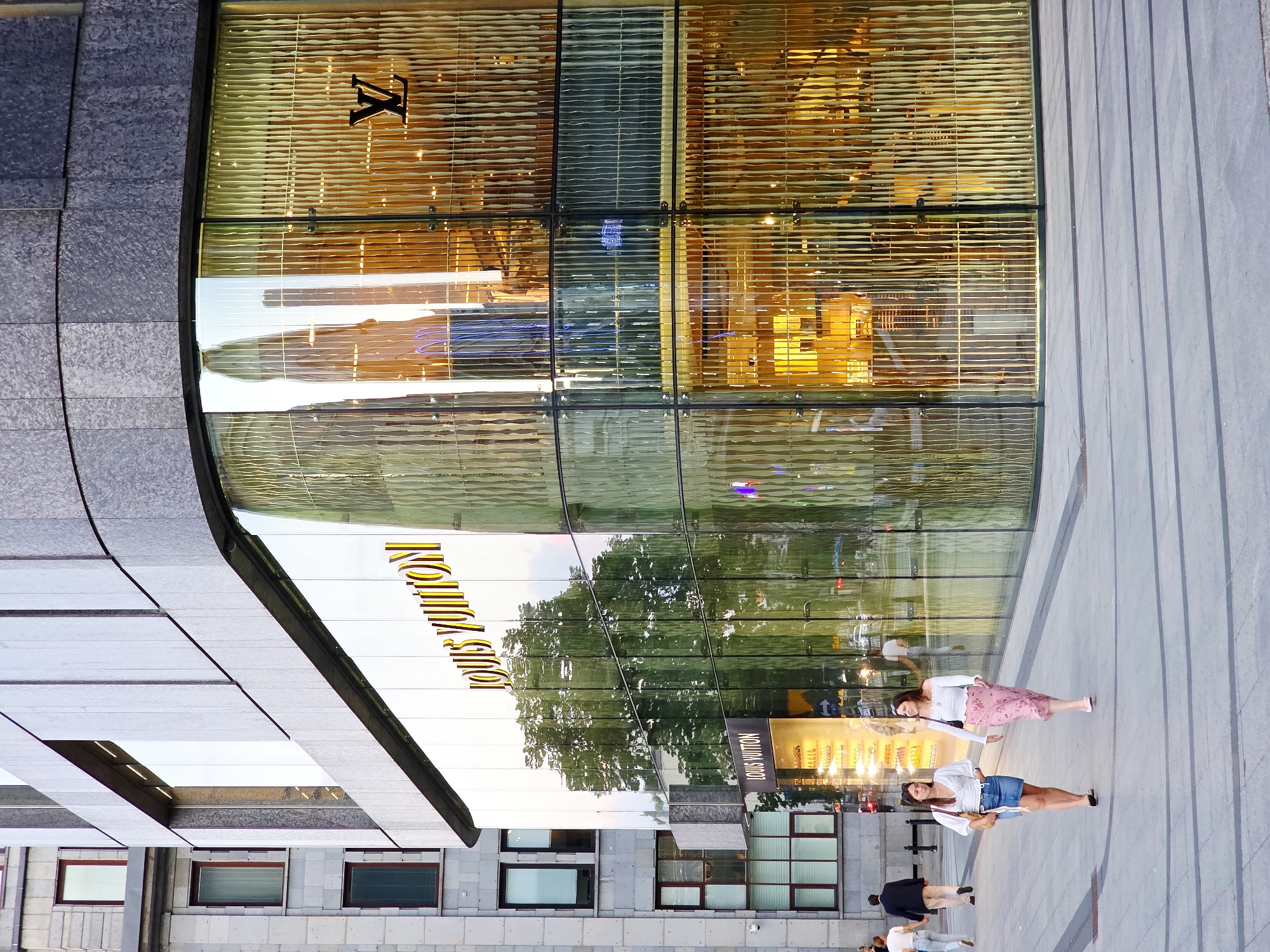 This is a full-shot, eye-level, outdoor photograph of the Louis Vuitton store at the corner of Nowy Świat Street and Aleje Jerozolimskie in Warsaw, Poland. The store is characterized by a large, curved glass facade with the Louis Vuitton logo prominently displayed in gold lettering. The interior of the store is illuminated with warm, yellow light, creating a reflection of the exterior environment, including trees and the sky, on the glass.
Two young women are walking on the sidewalk in front of the store. The woman on the left is wearing a white V-neck top, a denim skirt, and black flats. She is carrying a brown handbag. The woman on the right is wearing a white off-the-shoulder top and a pink floral skirt. She is also carrying a handbag. Other pedestrians are visible in the background, as well as traffic. The pavement in front of the store is made of grey tiles. The building next to the Louis Vuitton store is made of grey stone. The lighting suggests it is late afternoon or early evening.