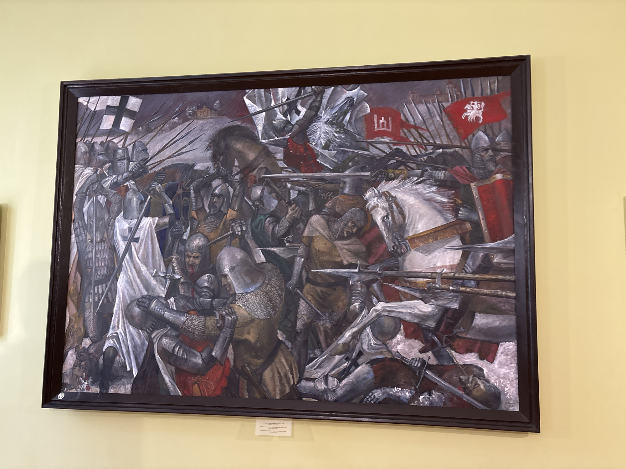 This is a photograph of a large, framed oil painting depicting a chaotic battle scene. The painting is mounted on a light yellow wall. A small white informational plaque is visible below the painting.
The painting itself is a detailed historical depiction of a medieval battle, likely a significant event in Belarusian history. It features numerous figures in armor, engaged in combat. Knights on horseback are prominently featured, some wielding swords and lances, while others are depicted fallen or injured. Flags with heraldic symbols are visible, including one with a white knight on a red background and another with a black cross on a white background. The artwork appears to be rendered in a realistic style with a dramatic, dark color palette, emphasizing the intensity and brutality of the conflict.
The caption on the informational plaque indicates that this artwork is located in the Hrodna State Museum of History and Archaeology in Hrodna, Belarus. The painting likely depicts a historical event that took place in or around the city of Hrodna.