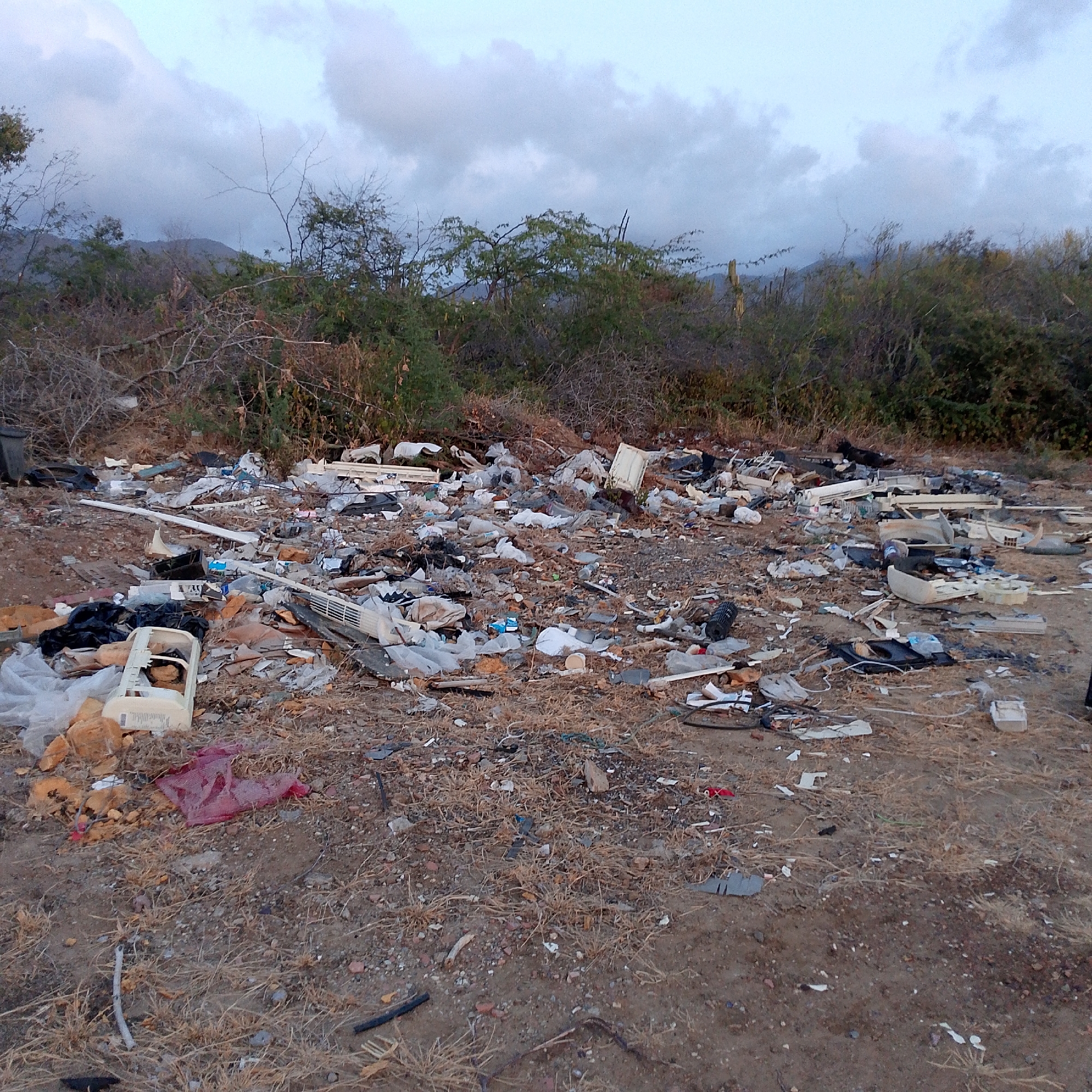 The image shows a large amount of discarded trash strewn across a barren landscape. The trash includes plastic bags, various containers, and broken appliances or electronic components. The ground is mostly dirt and sparse vegetation. In the background, there are trees and bushes, with hills or mountains visible in the distance. The sky is overcast. Given the location information, this scene is likely in or near Altagracia, Venezuela. There are no people visible in the image. The trash consists of white plastic containers and parts. There are black plastic bags mixed within the trash. The lighting indicates it is near dusk or dawn.