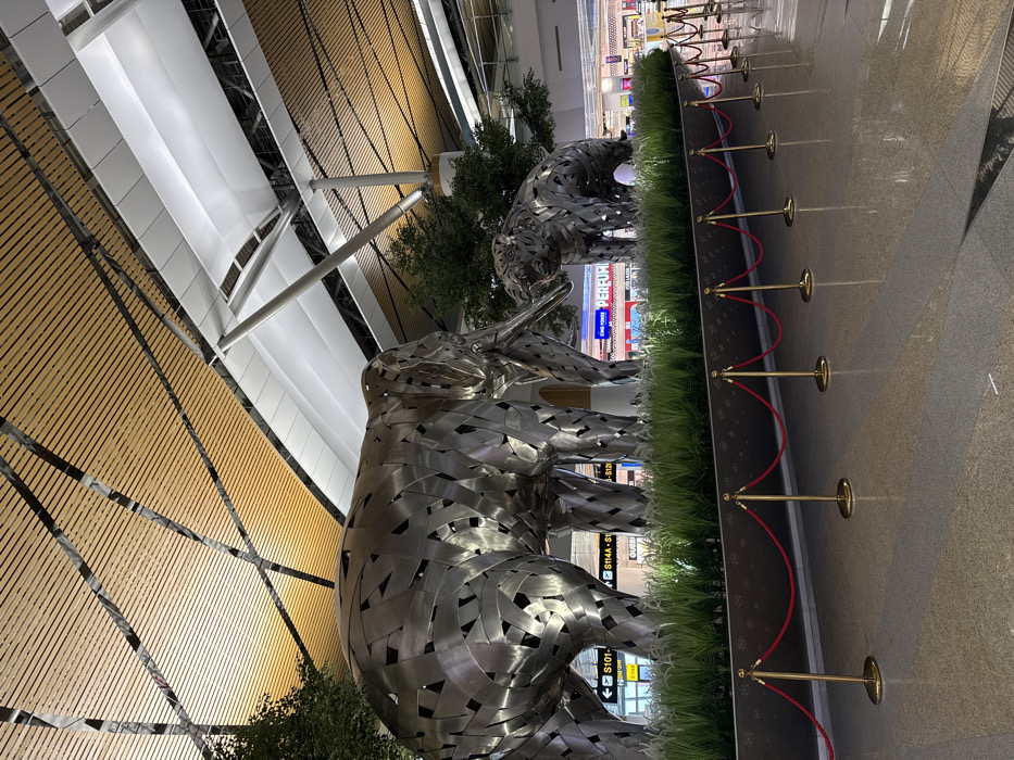 The scene depicts a modern indoor airport terminal in Racha Thewa Subdistrict, Thailand. In the foreground and central midground, two large, stylized animal sculptures constructed from segmented, reflective metallic panels are prominently displayed. The larger sculpture on the left is an elephant, shown standing. To its right, a smaller sculpture of a big cat, likely a tiger, also stands, with its mouth open as if roaring. Both sculptures are situated within a dark-colored planter containing artificial green grass and several small, artificial trees. This display area is cordoned off by a line of golden stanchions linked by red velvet ropes.
The polished, light-colored floor reflects the overhead lighting and portions of the display. In the background, airport signage, including a yellow directional sign with black text indicating gate numbers "S101-S104" and "S114A-S114B," is visible. Further back, shop fronts are discernible, one clearly marked "KING POWER PERFUME" in red lettering. The high ceiling features a complex design with parallel wooden slats interspersed with white, curvilinear fabric panels supported by large, diagonal metallic structural beams. The area is brightly illuminated by artificial overhead lighting. No individuals are visible within the frame.