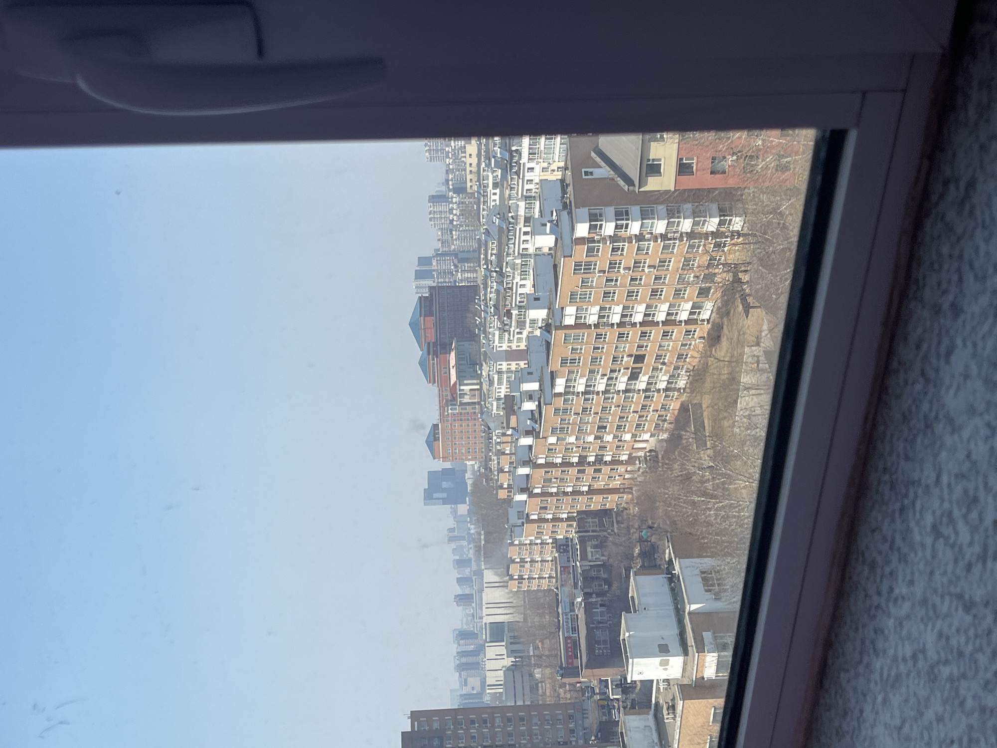The image displays a daytime aerial view of an urban landscape in Changchun City, China, observed through a window. A white window frame partially obscures the bottom and right edges of the scene, with a textured, light-grey surface (possibly a windowsill or floor) in the immediate foreground. The sky above is clear and light blue.
The midground is dominated by numerous multi-story residential buildings, typically five to seven stories in height, featuring beige or light brown facades with visible white window frames and enclosed balconies. Scattered among these structures are bare deciduous trees, indicating a late winter or early spring season. Various rooftops, some flat and white, others sloped and dark, are visible across the urban sprawl.
In the hazy background, a dense skyline of taller, modern high-rise buildings is discernible. Prominent background features include a large building with a reddish-brown facade and distinct blue-tiled traditional Chinese roofs situated towards the right, and a dark, angular skyscraper positioned towards the left. No individuals or overt actions are visible within the frame.