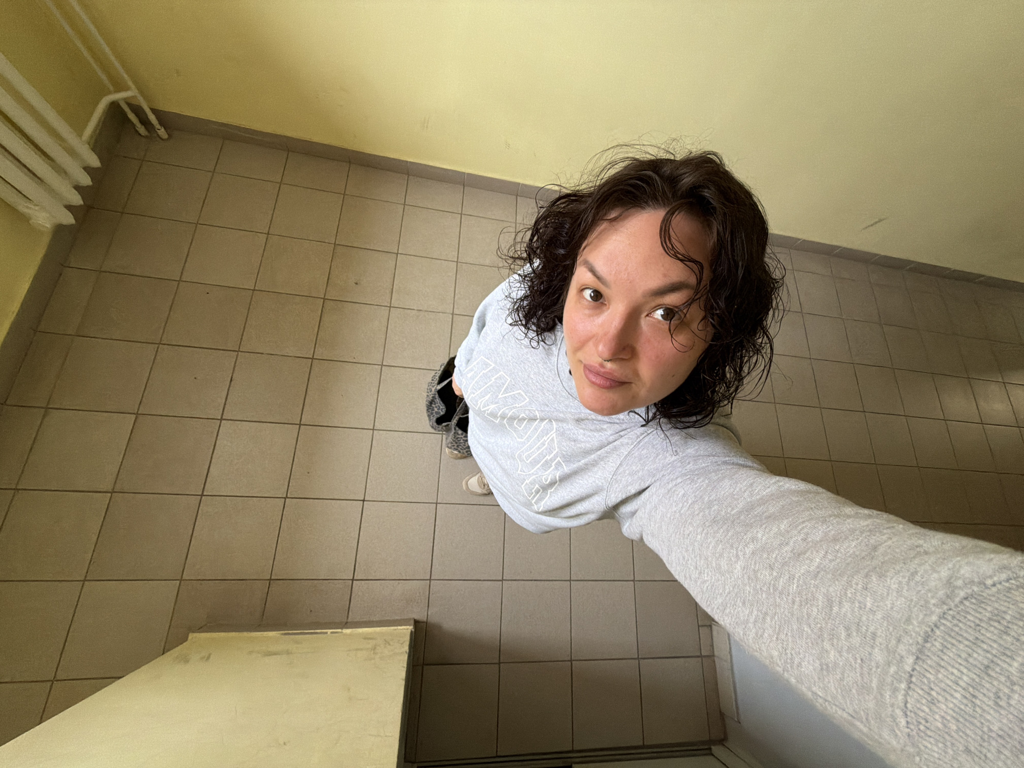 This is an indoor, high-angle, full-body selfie taken by a person with dark, curly hair and brown eyes. They are wearing a grey sweatshirt and grey sweatpants. The person is standing in a tiled hallway with beige floor tiles. To the left, there is a cream-colored wall and a part of what appears to be a doorframe or partition. To the right, there is a pale yellow wall. Along the top of the yellow wall, white pipes or a radiator system are visible. The image does not contain any information that can be used to determine the location in the city of Chaikovsky, Russia.