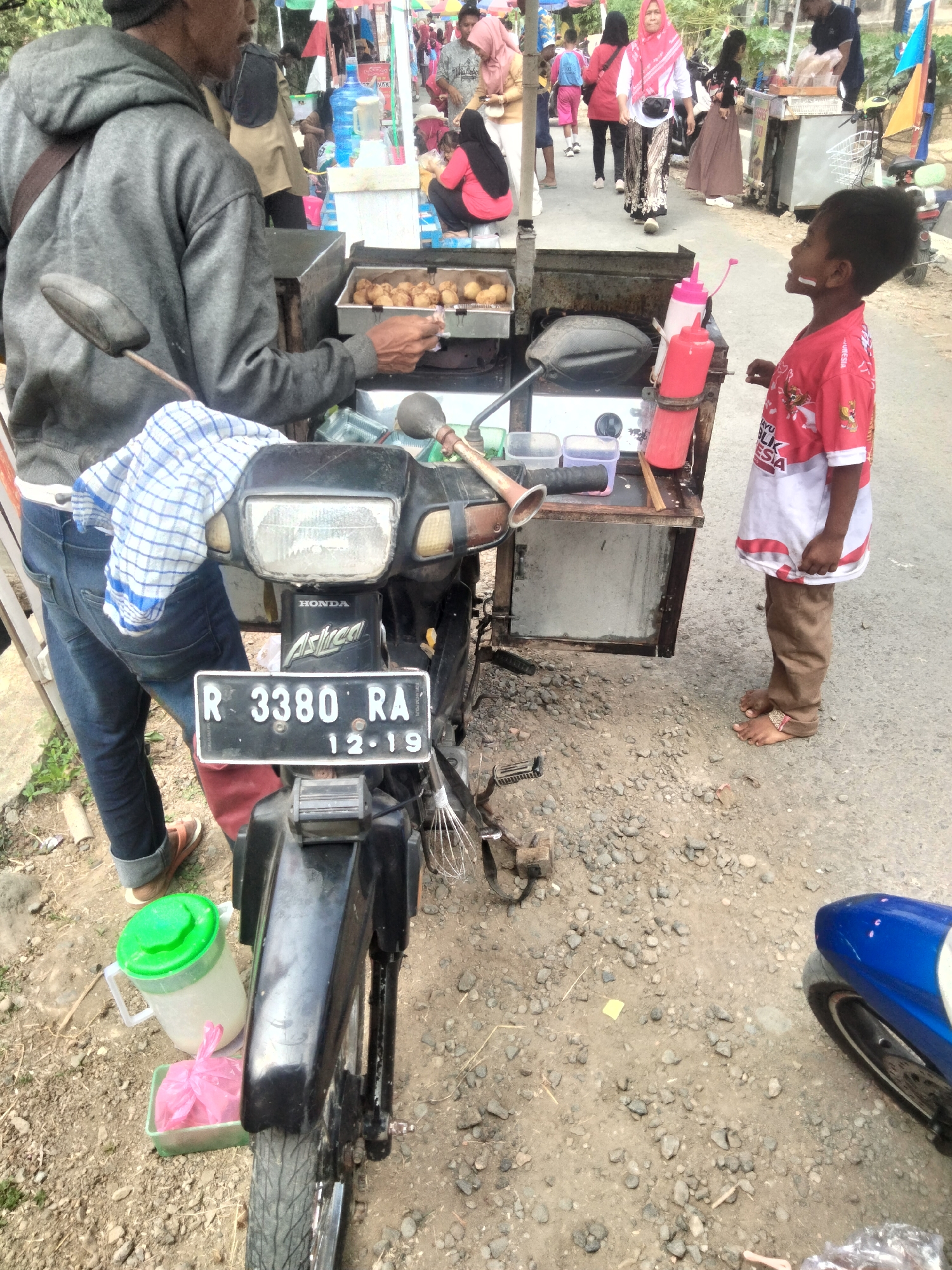 In Ketanggung, Indonesia, a street vendor is preparing food on a cart attached to a motorcycle. The vendor, a man wearing a grey hooded sweatshirt and blue jeans, is standing behind the cart, which is equipped with a cooking apparatus. He is holding a food item and appears to be in the process of serving it. A young boy, dressed in a red and white shirt with Indonesian text and brown pants, stands on the right side of the cart, looking up at the vendor. The cart holds various containers, including condiment bottles and a large water jug with a green lid. In the foreground, a motorcycle is parked, with its license plate visible as "R 3380 RA 12-19." The ground is covered with gravel and dirt. In the background, other people and market stalls suggest a busy public area.