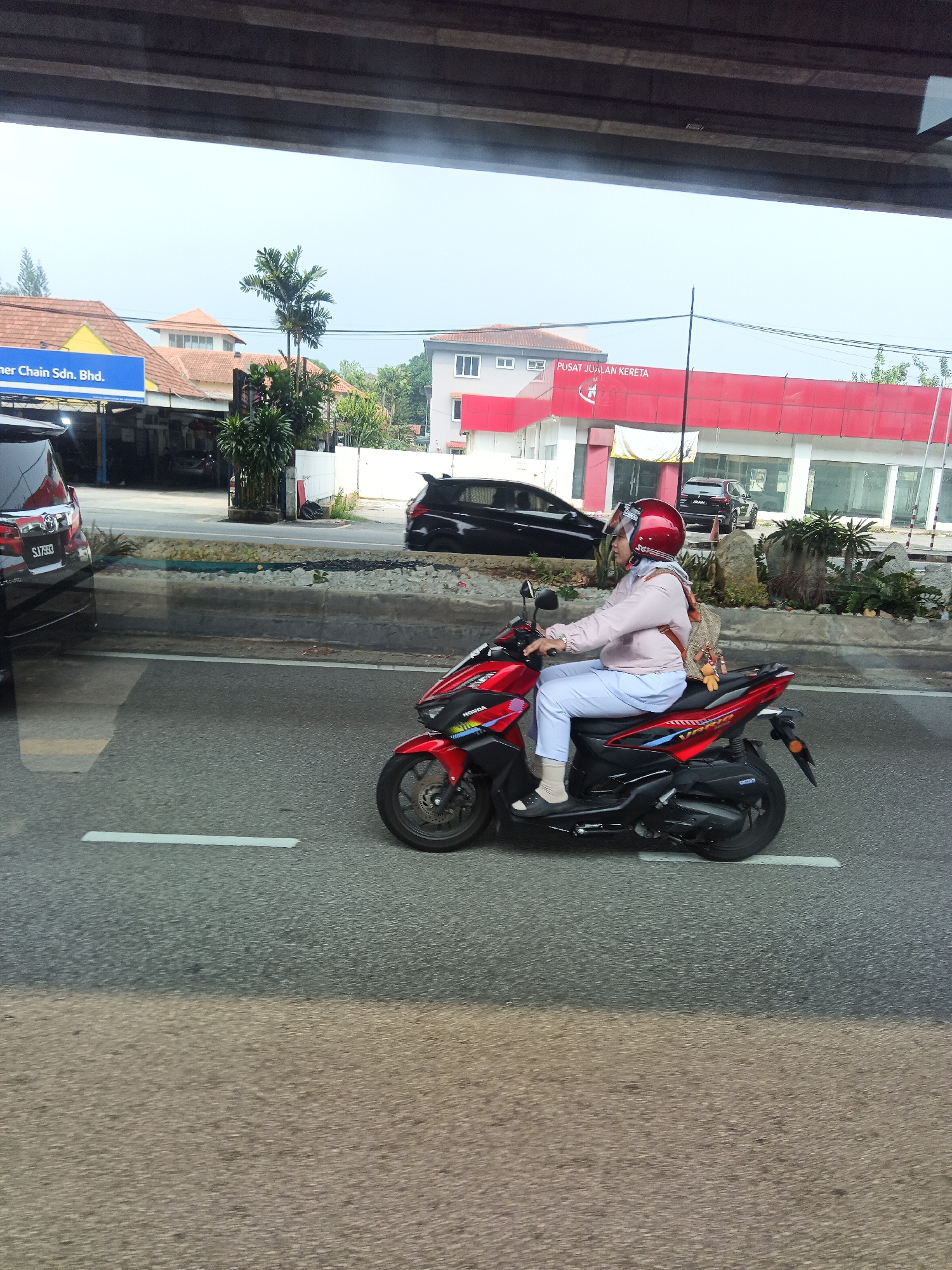 The image shows a woman riding a red scooter on a road. She is wearing a pink jacket, light blue pants, white socks, and a red helmet. She has a backpack on her shoulders. The scooter has "HONDA" written on it. In the background, there is a building with a red facade that says "PUSAT JUALAN KERETA" (Car Sales Center). There are also other cars and buildings visible. The image appears to be taken from another vehicle, as indicated by the reflection on the left side of the image and the blurred foreground. The scene is set in an urban environment, possibly a commercial area in Kuala Lumpur, Malaysia, given the presence of Malaysian script on the building and the typical urban architecture.
