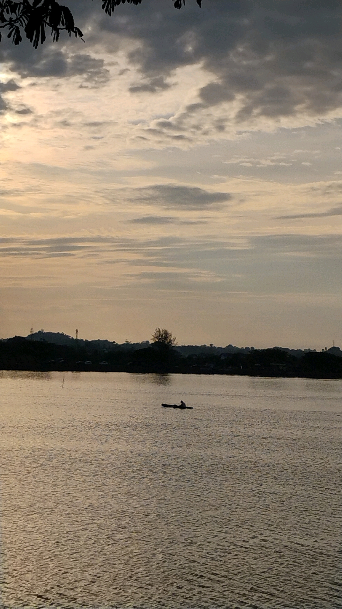 An individual is silhouetted in a kayak on the surface of a body of water, likely a river or inlet, in Lhokseumawe, Indonesia. The sky is overcast with streaks of light indicating sunrise or sunset, casting a soft, muted light on the scene. In the background, a dark, silhouetted treeline marks the shore, with faint outlines of structures and possibly some vehicles visible. The water's surface is rippled, reflecting the subdued colors of the sky. Tree branches, silhouetted against the sky, frame the top left corner of the image.
