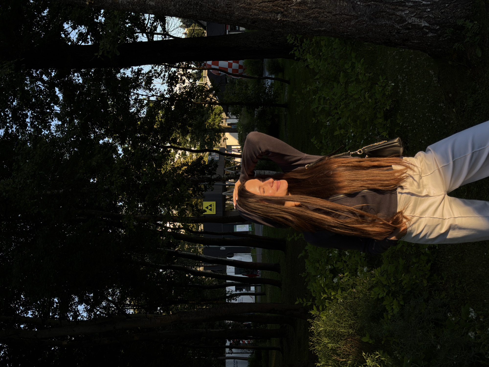 In the city of Odintsovo, Russia, a young woman with long, brown hair stands in a park at golden hour, her body angled to the right, her left arm bent with her hand resting on the back of her head. She wears a dark long-sleeved top and light-colored pants, and a black purse with a gold chain hangs from her shoulder. The low sun casts a warm glow on her face and illuminates the foliage around her. Behind her, a line of trees with dark trunks creates a dappled shade effect, and the background reveals a building with a yellow triangular logo and the word "FISCHER" printed on it. Further to the right, a large tree trunk is visible, its bark textured and dark. The ground is covered in green grass and low-lying plants.