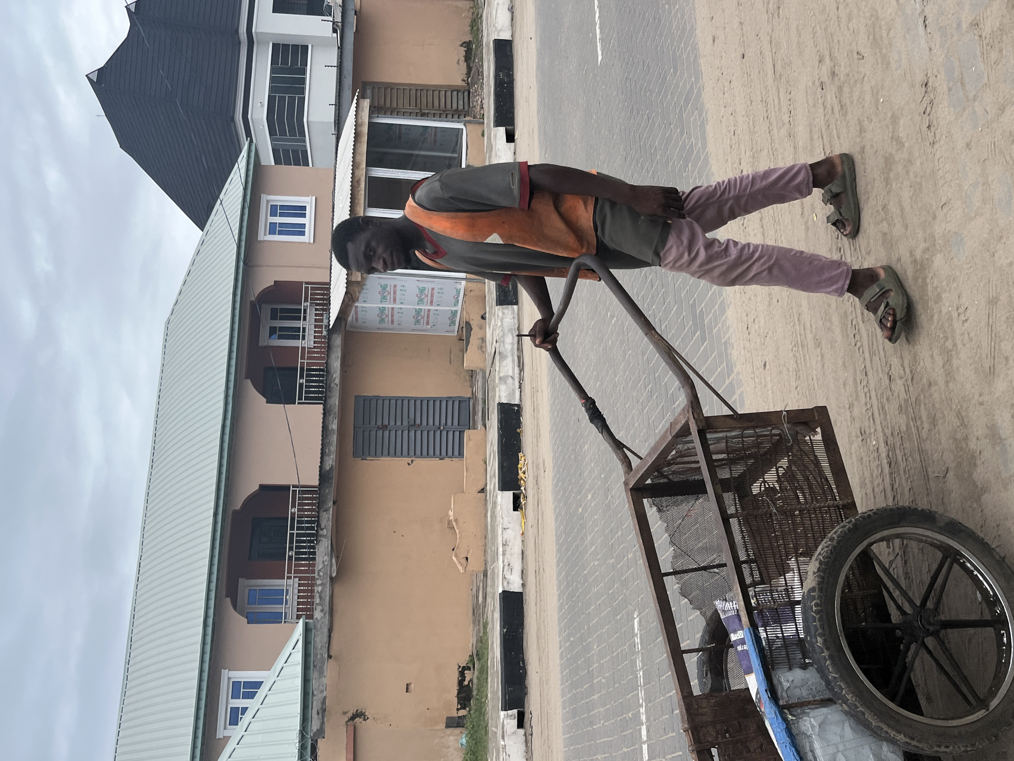 A man stands on a brick-paved street in Badagry, Nigeria, pulling a metal cart. He wears a muted orange vest over a gray t-shirt, pink pants, and green sandals. The cart contains a large tire and other indiscernible objects. Buildings with multiple stories, peach-colored facades, white trim, and dark gray roofs line the street. A black and white painted curb is visible. The sky is overcast.