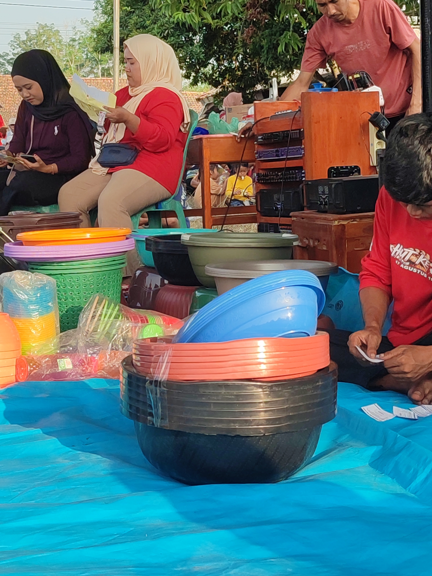 A wide shot captures an outdoor market scene in Nusa Jaya, Indonesia, dominated by a bright blue tarp spread across the ground. In the foreground, a stack of black and orange plastic bowls is prominently displayed. To the left, a collection of smaller plastic containers in various colors, including green, blue, yellow, and orange, are piled up, some wrapped in plastic.
In the mid-ground, two women are seated. The woman on the left, wearing a black hijab and a maroon top, is looking down at a mobile phone in her hand. Beside her, a woman in a light beige hijab and a red long-sleeved shirt is holding up a yellow document, appearing to read it. She is seated on a green plastic chair. Behind them, a man wearing a reddish-pink t-shirt is attending to electronic equipment on a wooden stand. The equipment appears to be audio-related, with amplifiers and other devices visible. Trees with green foliage form the backdrop of the scene. A building with a terracotta-tiled roof is partially visible in the upper background. The ground beneath the tarp appears to be dirt or sand.
