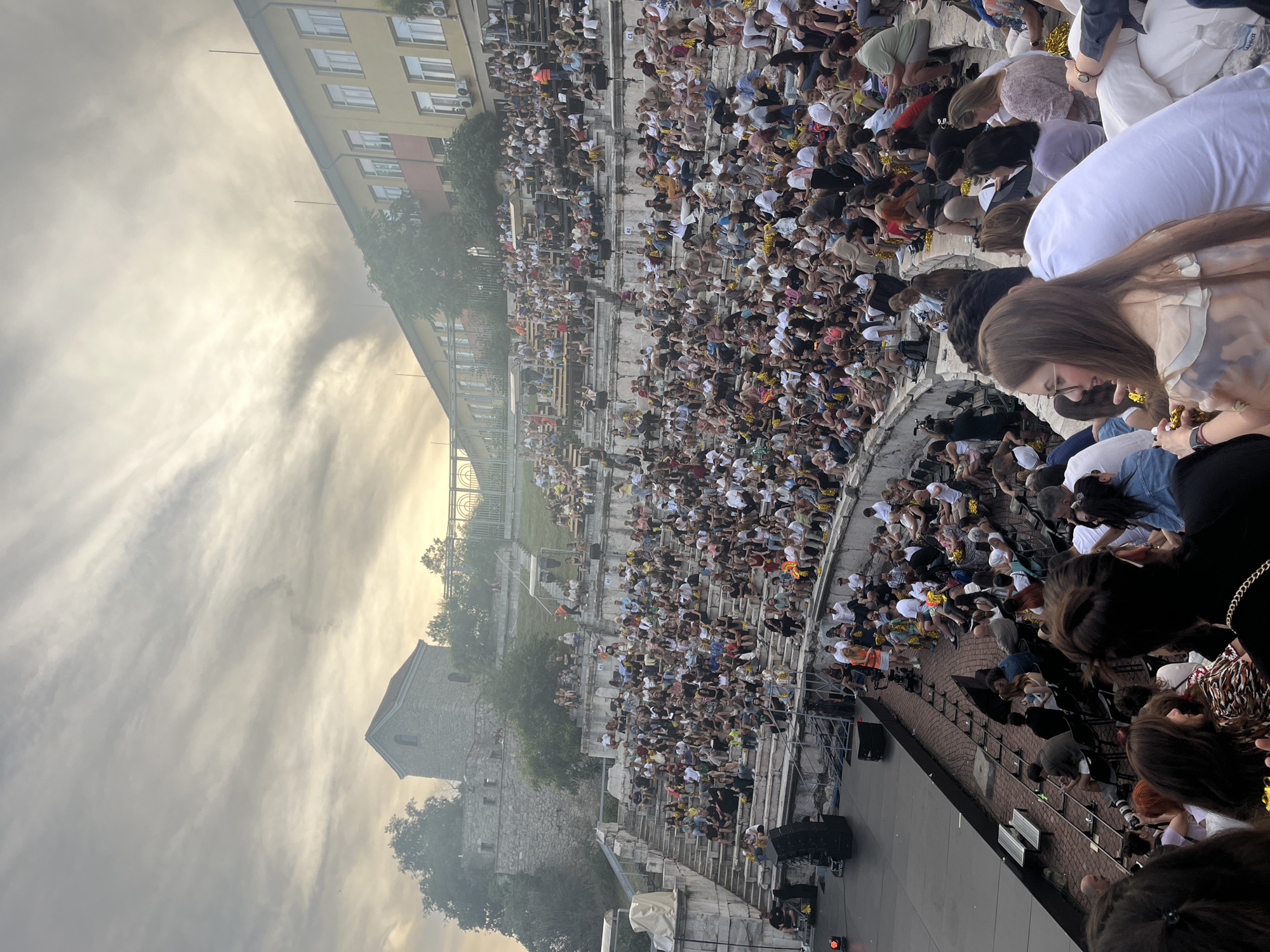 An outdoor amphitheater is filled with a large audience seated on stone steps. The audience is a diverse group of people of various ages, dressed in casual summer clothing. Many are holding yellow pompoms. In the background, there is a stage with speakers and lighting equipment. Behind the amphitheater, a yellow building with many windows stands against a cloudy sky. To the left of the building, there is a stone structure that appears to be part of an ancient ruin, with trees growing around it. The lighting suggests it is late afternoon or early evening.