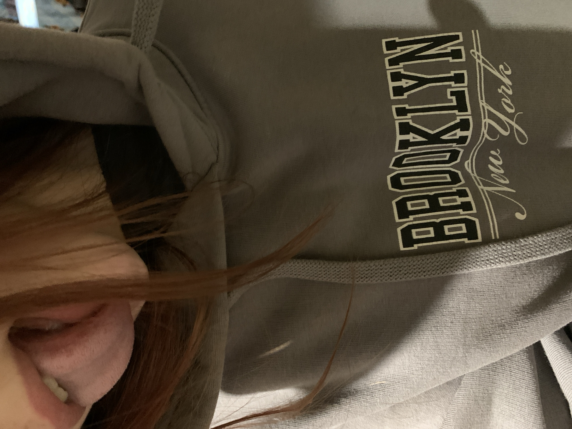 The image shows a close-up of a person wearing a gray hoodie with the words "BROOKLYN New York" printed on it. The person's mouth is visible with their tongue sticking out. The background is out of focus and indistinct. The city and country of Фоки, Russia are not apparent in the image.