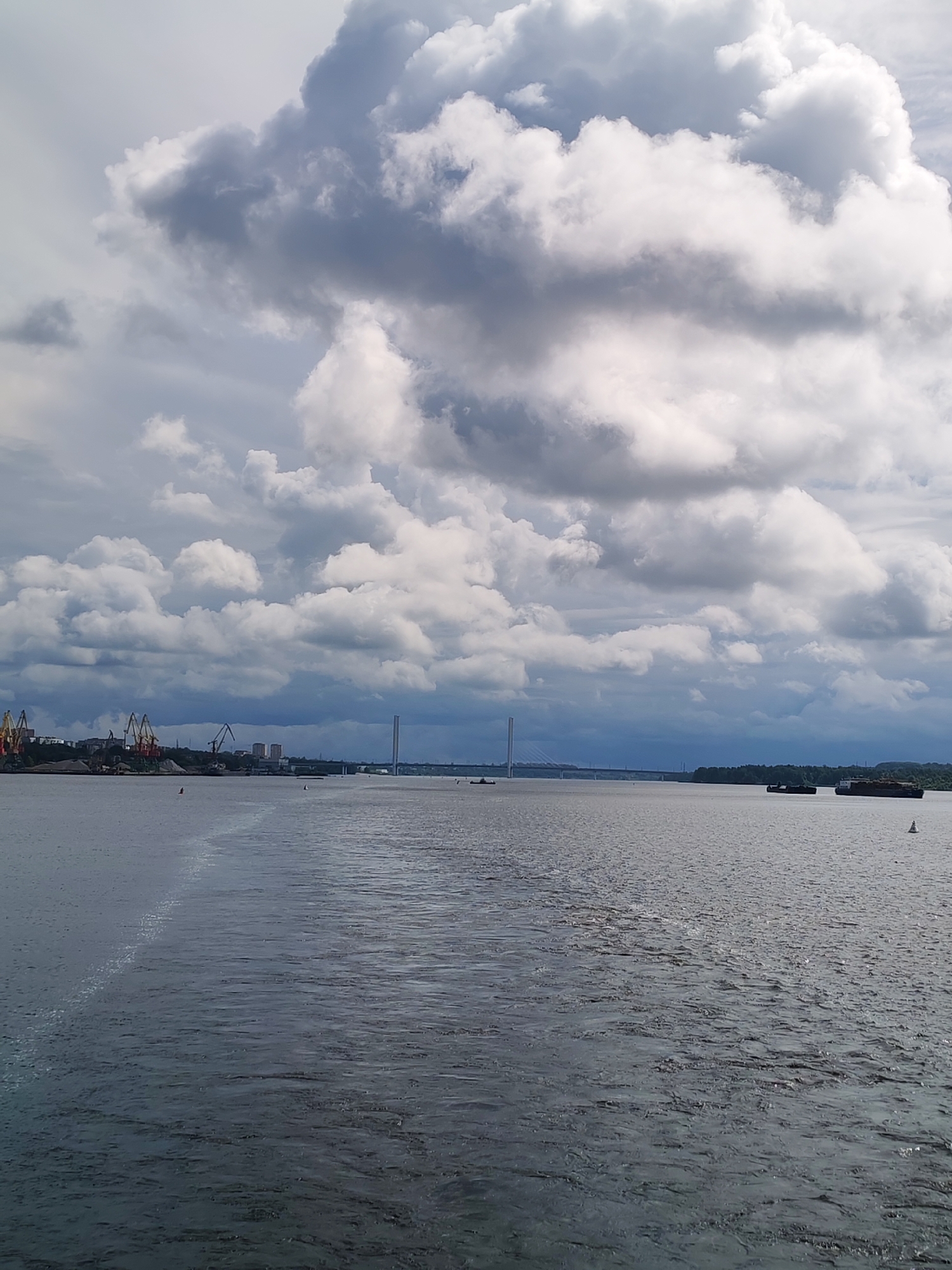 The image depicts a wide body of water, likely a river or bay, with a shoreline visible in the distance. The sky is filled with dramatic, heavy clouds, some dark and foreboding, others bright and illuminated, suggesting a turbulent weather system.
On the left side of the distant shoreline, industrial cranes and piles of material indicate a port or industrial area. Further along the shore, there are buildings and what appears to be a large suspension bridge spanning the water. To the right, more of the shoreline is covered in trees, and several barges or cargo ships are present on the water, with buoys marking the waterway. The water itself has a textured surface, reflecting the light and showing patterns from movement or wind.
This scene is consistent with the Sheksna River in Cherepovets, Russia, specifically the view from a vessel approaching or departing the city's port, with the Northern Bridge (Severny Most) visible in the distance. There are no people visible in the image.