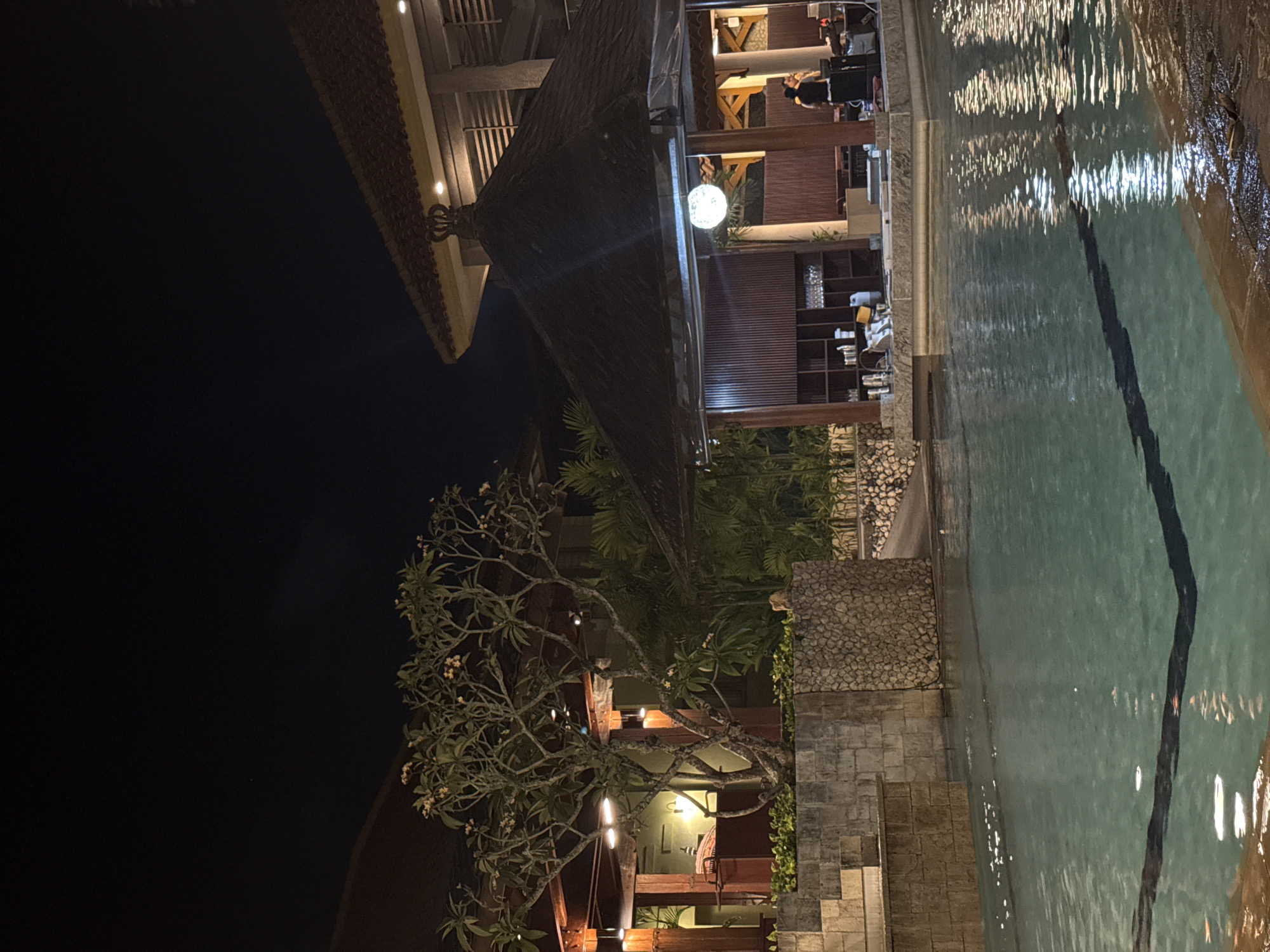 This image depicts a nighttime scene at a resort in Kuta Selatan, Indonesia, likely a poolside bar or restaurant area. In the foreground, a swimming pool with turquoise water is illuminated by reflections from overhead lights. The pool has a dark blue line marking its edge. To the left, a stone wall structure with steps leads up from the pool, and a tree with sparse foliage and small flowers is planted in a stone planter.
Behind the pool, there is a covered outdoor dining or bar area with a dark, thatched roof supported by wooden posts. Inside this structure, shelves are stocked with glasses, and tables and chairs are set up. A few people are visible; one person, dressed in dark clothing, appears to be standing behind a counter or bar, possibly a staff member. Another person can be seen further back within the covered area. The background features more foliage and resort architecture, with wooden beams and tiled roofs visible. The overall ambiance is of a tropical resort at night, with warm lighting contributing to a relaxed atmosphere.