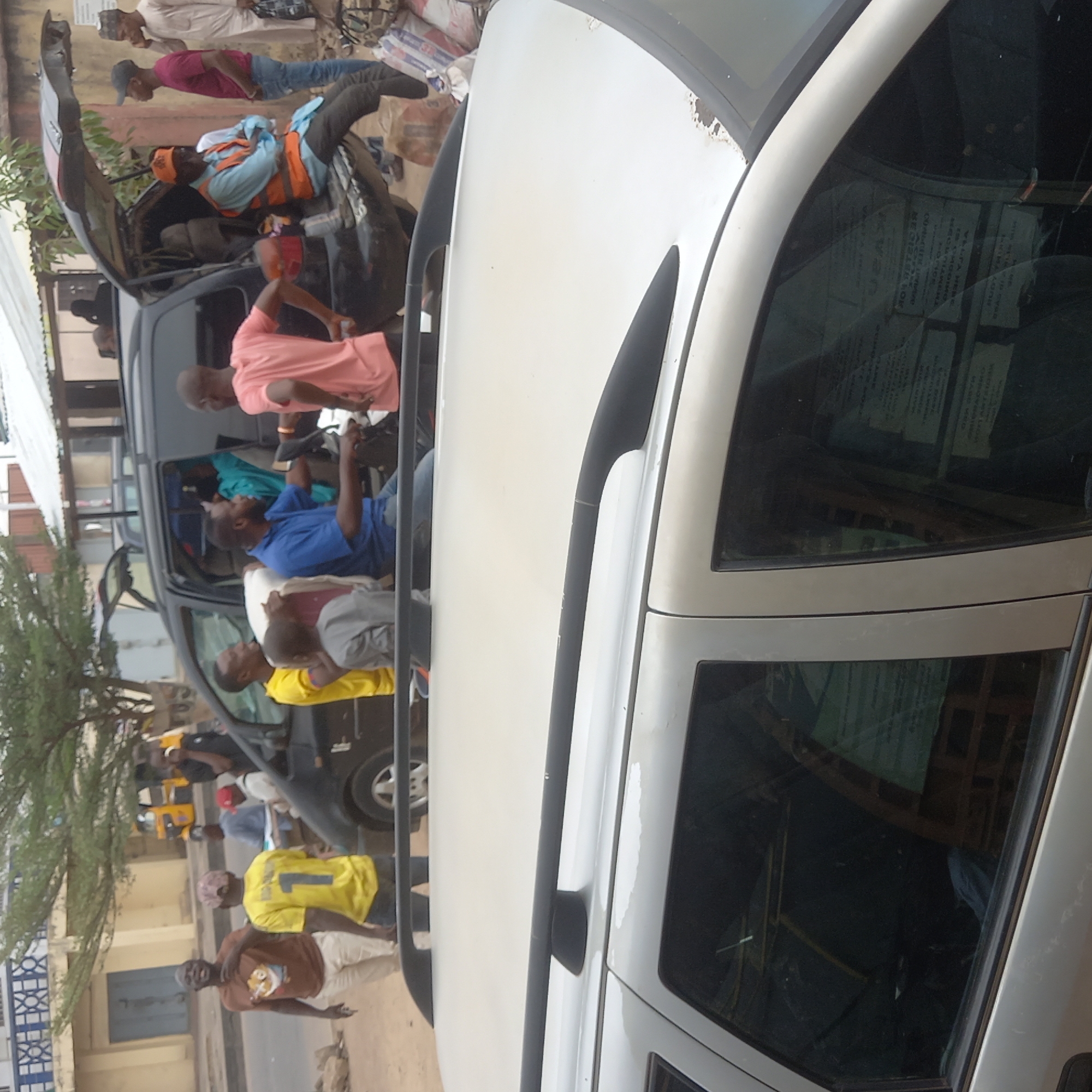 The image shows two vehicles in Dadi Riba, Nigeria. A black van is filled with passengers. One man in the van wears an orange vest. People are standing outside the van. A white car is in the foreground. The image is taken in a street setting.