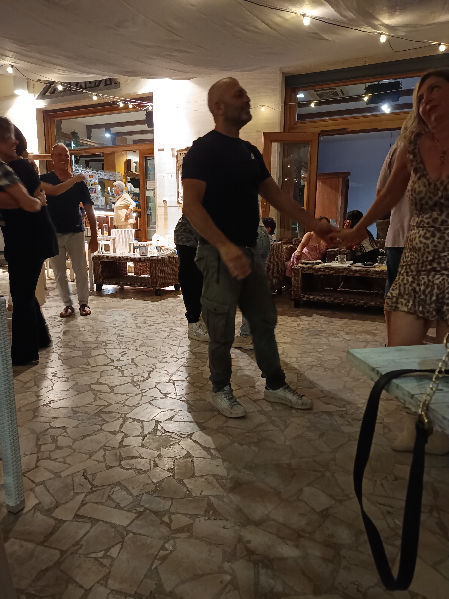 The image depicts an outdoor scene at night, possibly a restaurant or bar patio in Pineto, Italy. String lights are visible overhead. Several people are present, some of whom appear to be dancing or socializing. In the foreground, a bald man in a black t-shirt and grey cargo pants is dancing with a woman in a leopard-print dress. To the left, a woman in a black dress is also dancing, and a man in a blue t-shirt and white pants is standing nearby. In the background, other individuals are seated at tables or moving around. The ground is paved with irregular stone tiles.