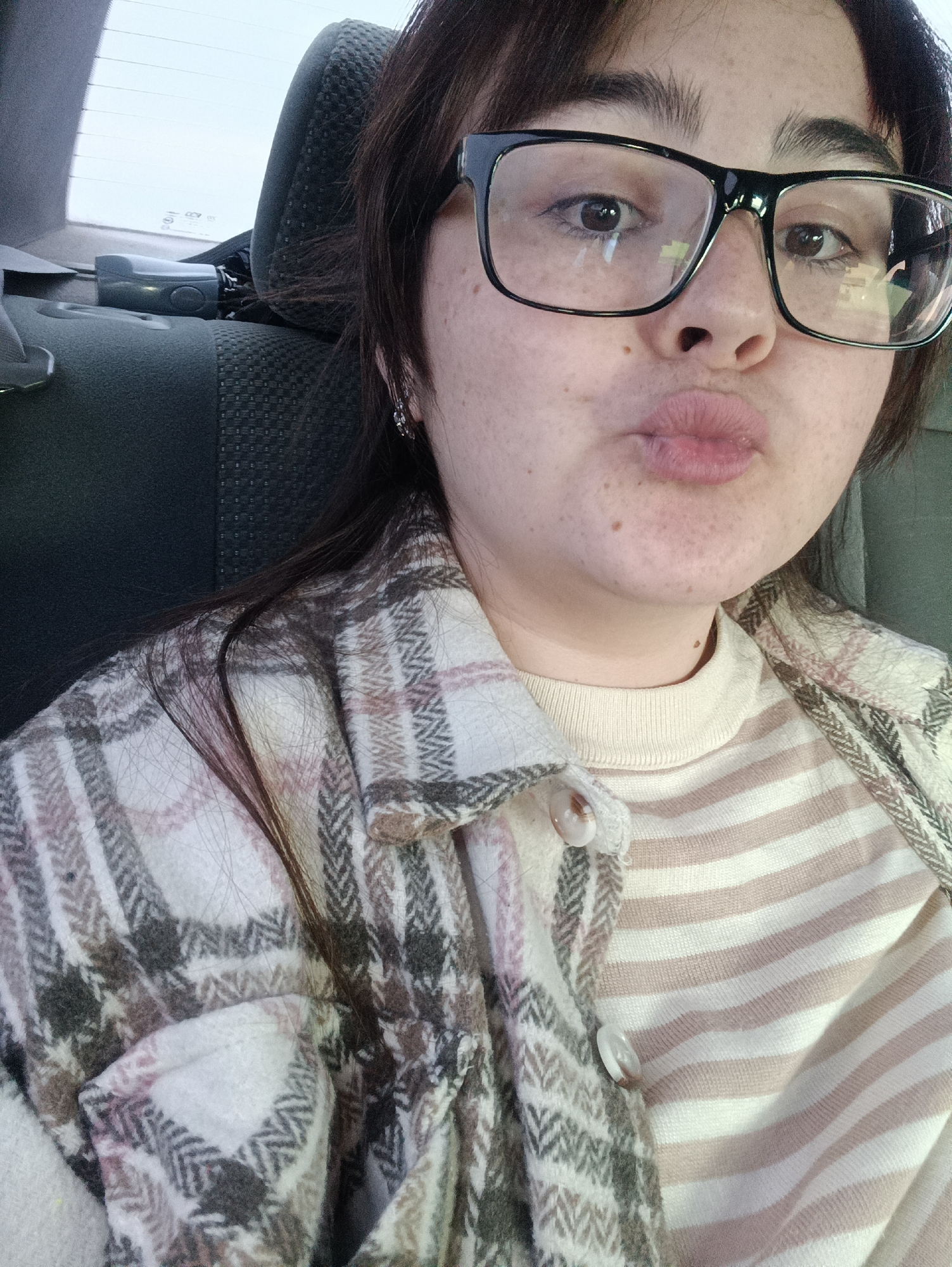 A woman is sitting in the back seat of a car. She is wearing large, black-framed glasses. She is wearing a striped shirt and a plaid jacket. She has dark hair and her lips are pursed. The car seat is gray. The photo was likely taken in Chaikovsky, Russia, based on a presumed association with the city name.