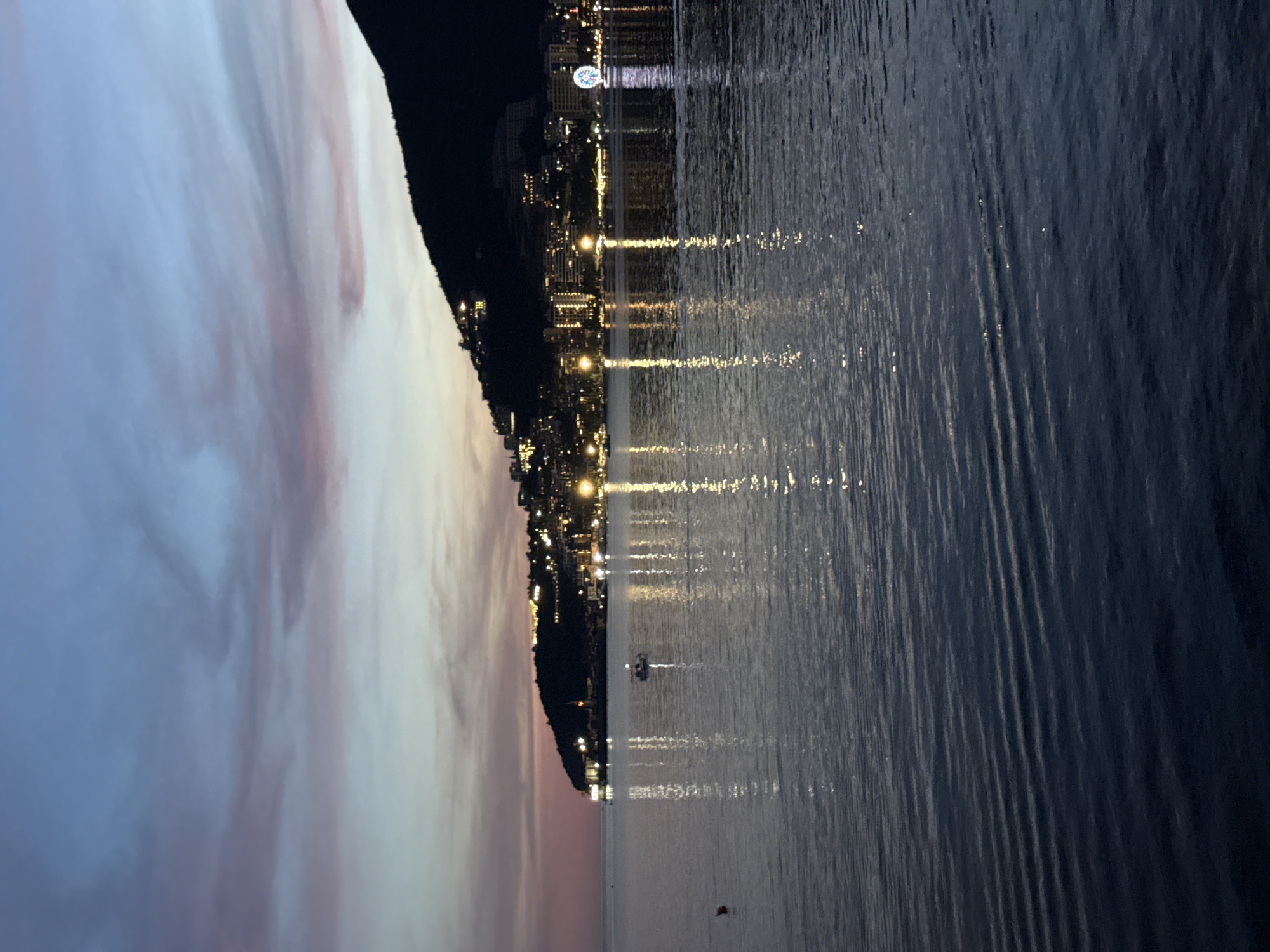 The image shows a twilight view of Budva, Montenegro, with the city lights reflecting on the calm sea. A dark, tree-covered hill dominates the right side of the frame, sloping down towards the water. Along the coast, buildings are illuminated with warm yellow lights, creating a vibrant contrast against the darkening sky. In the distance, a Ferris wheel is visible, a prominent landmark of Budva's promenade. The sky exhibits hues of deep blue and soft pink, with streaks of clouds indicating the end of daylight. The water's surface is smooth, with gentle ripples that distort the reflections of the city lights into elongated streaks. A small boat with two occupants is visible in the middle ground, gently moving on the water. A red buoy is also present in the foreground on the left. The overall scene is serene and picturesque, capturing the atmospheric beauty of Budva at dusk.