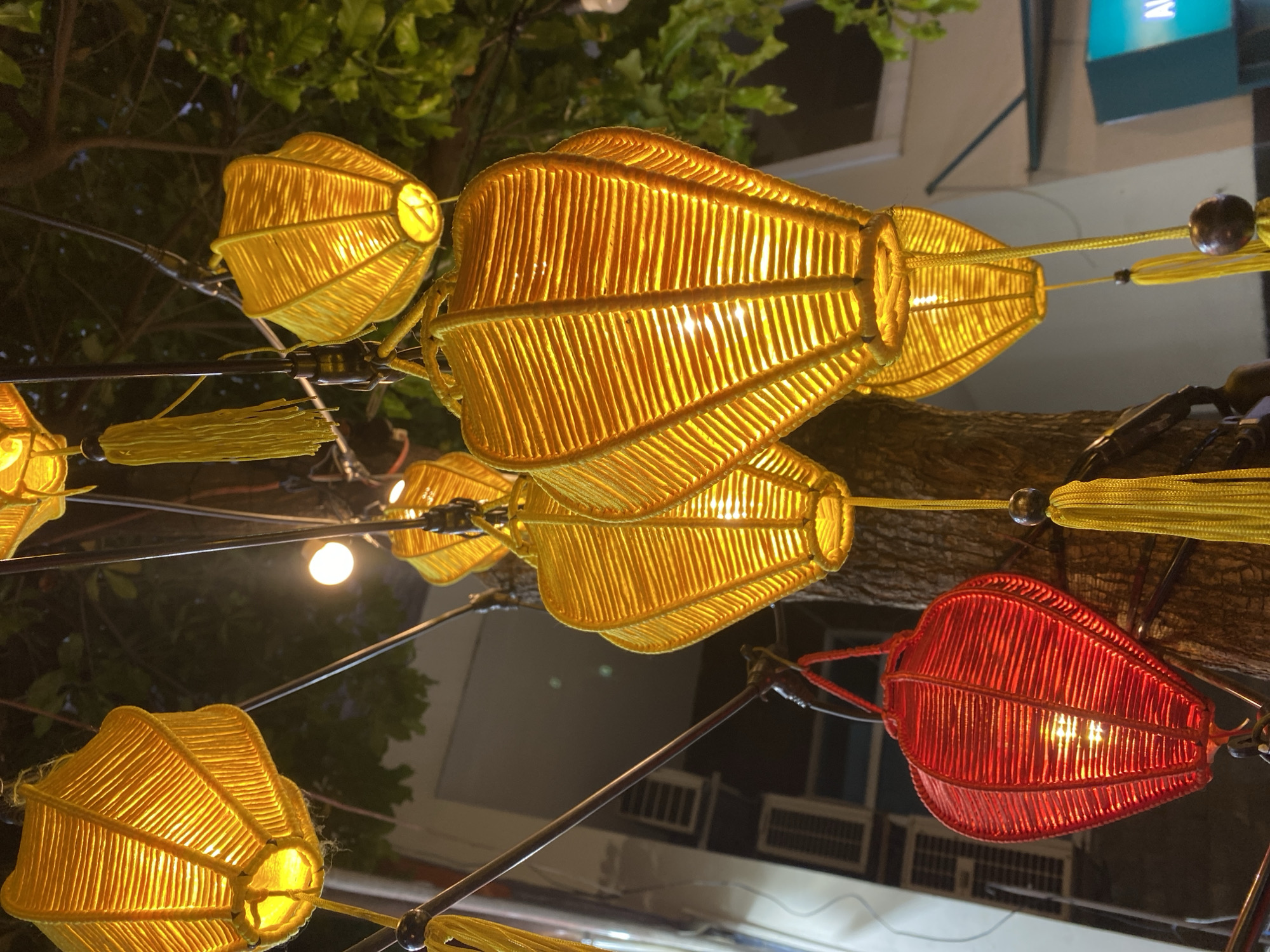 A close-up, low-angle shot captures a cluster of brightly lit, heart-shaped lanterns hanging from a tree in the city of Đà Nẵng, Vietnam. The lanterns, made of woven yellow and red threads, cast a warm glow. Several yellow lanterns are visible in the foreground and middle ground, one larger red lantern is in the lower left, and another yellow lantern is in the upper left. All the lanterns have a textured, ribbed appearance and are illuminated from within. Yellow tassels hang from some of the lanterns. The background is dark, with hints of greenery from the tree's foliage and a blurry glimpse of a building with air conditioning units. The image appears to be taken at dusk or night.