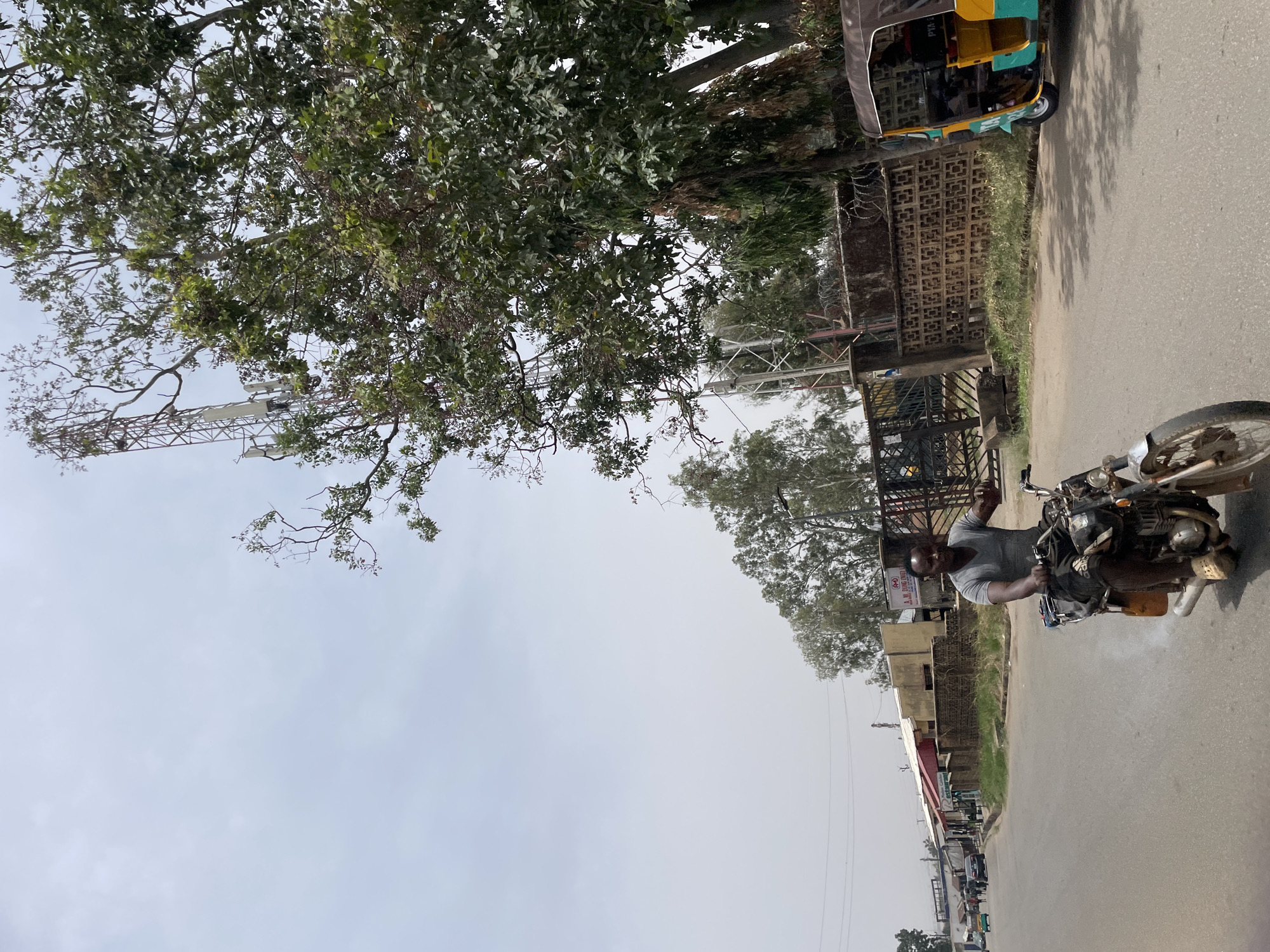 A street scene in Jos, Nigeria, shows a man on a motorcycle in the right foreground, traveling on a paved road. He is wearing a t-shirt and is holding one hand up as if waving. In the upper right corner, a vehicle similar to a tuk-tuk is parked near a building with decorative brickwork and a metal gate. On the left side, a cellular tower rises behind a large tree, partially obscured by foliage. Further down the road on the left, buildings and other vehicles suggest a commercial area. The sky is overcast, casting a muted light on the scene.
