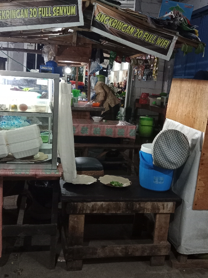 This image depicts an outdoor food stall, identified by banners as "Angkringan 20," located in the city of Turunsih, Indonesia, during nighttime. The stall operates under a provisional wooden roof structure adorned with two banners: "ANGKRINGAN 20 FULL SENYUM" and "ANGKRINGAN 20 FULL WIFI."

In the mid-ground, an individual, appearing to be an adult, is partially visible from the back. They wear a head covering and a brown patterned garment, positioned behind a counter covered with a pink and white geometric patterned tablecloth, engaged in an activity consistent with food preparation or service. A white bowl is present on this counter. To the left, a glass display case exhibits various food items, including an orange, spherical item and white rectangular items on the upper shelf, and stacked white disposable bowls on the lower shelf, next to a blue plastic bag.

In the foreground, a dark wooden table supports two light-colored, wavy-edged plates; one contains green leafy herbs. A blue plastic cooler with a grey and white checkered cap on its lid is situated on the right side of this table. Green plastic holders containing green straws are visible on the main counter. The environment includes additional green buckets and various hanging items in the background, suggesting inventory or general merchandise. Artificial lighting illuminates the scene, indicating the time of day is after dusk.