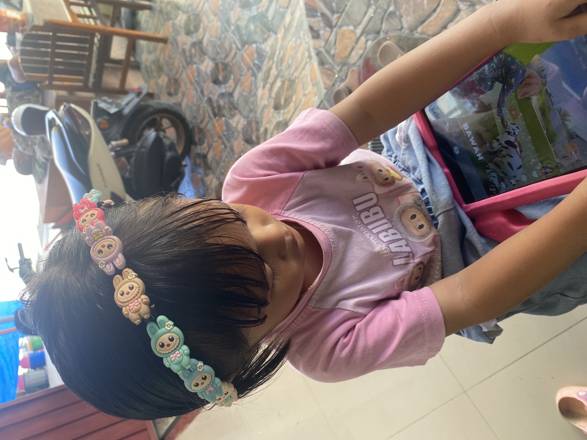 A young child with dark hair wears a pink shirt with a cartoon graphic and a pink headband adorned with cartoon bunny charms. The child is looking down at a pink tablet computer that displays a video. The child is seated on what appears to be a blue denim garment. The background shows a tiled floor, a motorcycle, and wooden furniture. The image is taken outdoors, likely in a residential area or an open-air space in Ta', Indonesia, as suggested by the style of the flooring and furniture.