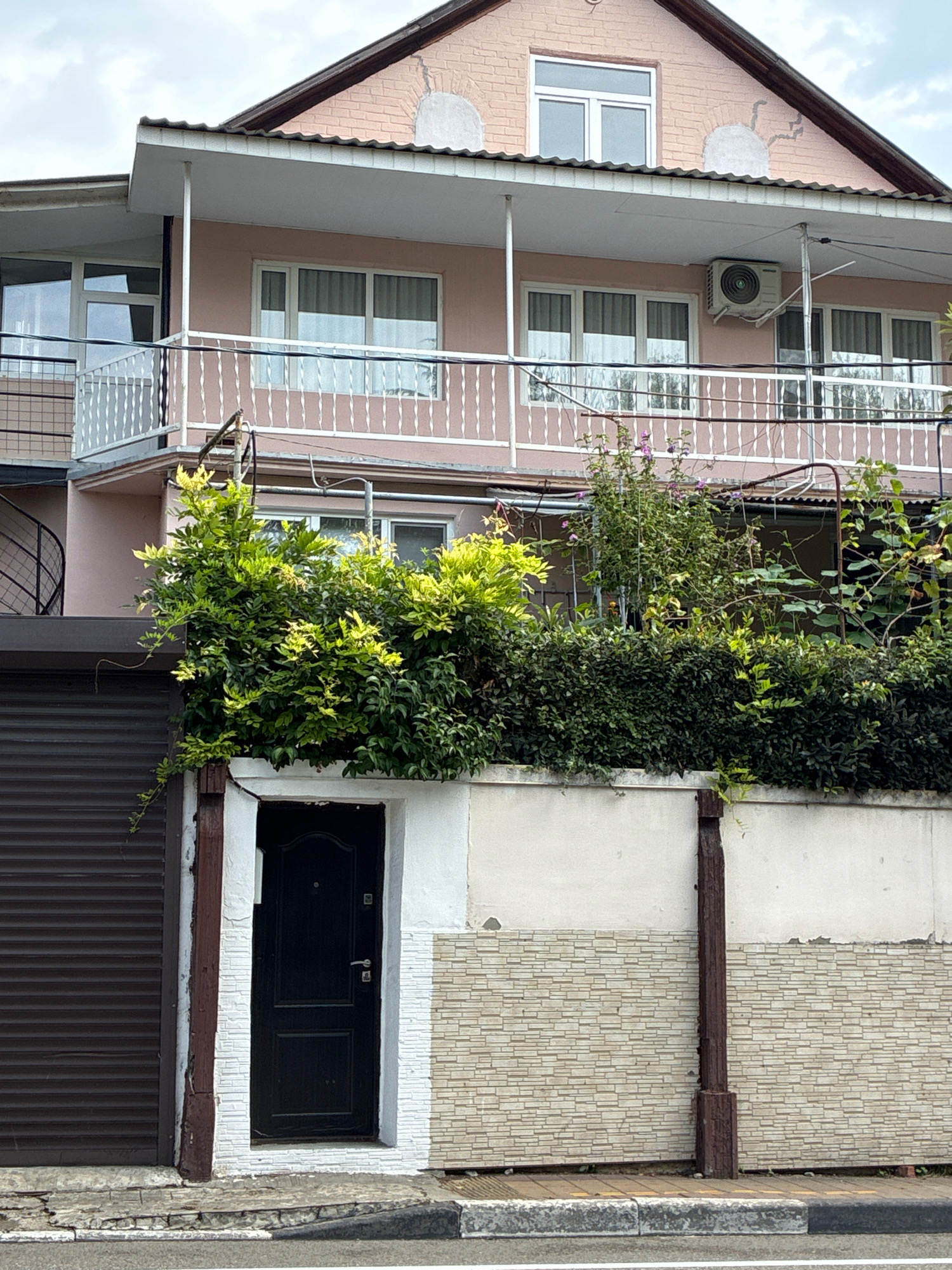 This is a daytime, eye-level, medium shot of a building in Sochi, Russia. The building is a two-story structure with a pinkish-peach facade. The lower story has a dark brown metal roll-up garage door on the left and a black door framed by white and light brown tiled stone on the right. A white fence with decorative spindles runs along the second-story balcony, which overlooks lush green foliage. The roof is tiled and slopes downwards. An air conditioning unit is mounted on the exterior wall of the upper story. The scene includes a sidewalk and a street in the foreground. The sky is overcast with scattered clouds.