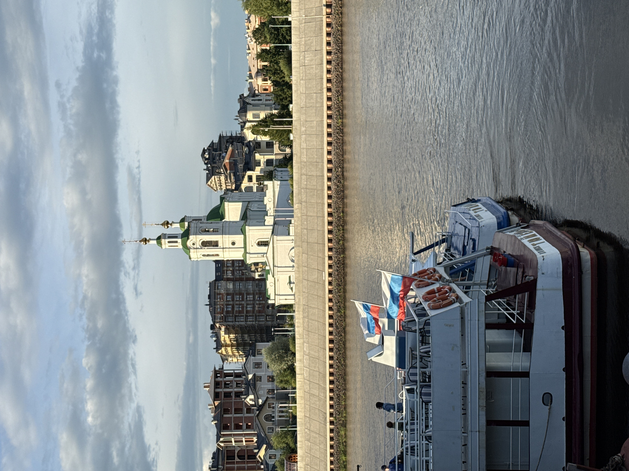 This is a photograph taken from a body of water, looking towards a city skyline. In the foreground, there are two boats docked. The boat on the left is larger and appears to be a passenger vessel with multiple levels and windows. It has a red and white hull and is adorned with two Russian flags. The boat on the right is smaller and blue and white.
In the middle ground, there is a reinforced concrete embankment with a row of cylindrical structures along its base. Behind the embankment, the city skyline is visible, dominated by a white church with a green roof and two prominent green domes topped with crosses. Various other buildings, including residential structures with brick and light-colored facades, are situated to the left and right of the church. Some of these buildings appear to be under construction, with scaffolding visible. Trees and lampposts are interspersed among the buildings.
The sky above is partly cloudy, with the sunlight casting a warm glow on the scene. The water in the foreground is calm, reflecting the sky and the surrounding structures. The overall impression is a serene, urban waterfront scene.
The image depicts the waterfront of Tyumen, Russia, with the Transfiguration Cathedral in the background.