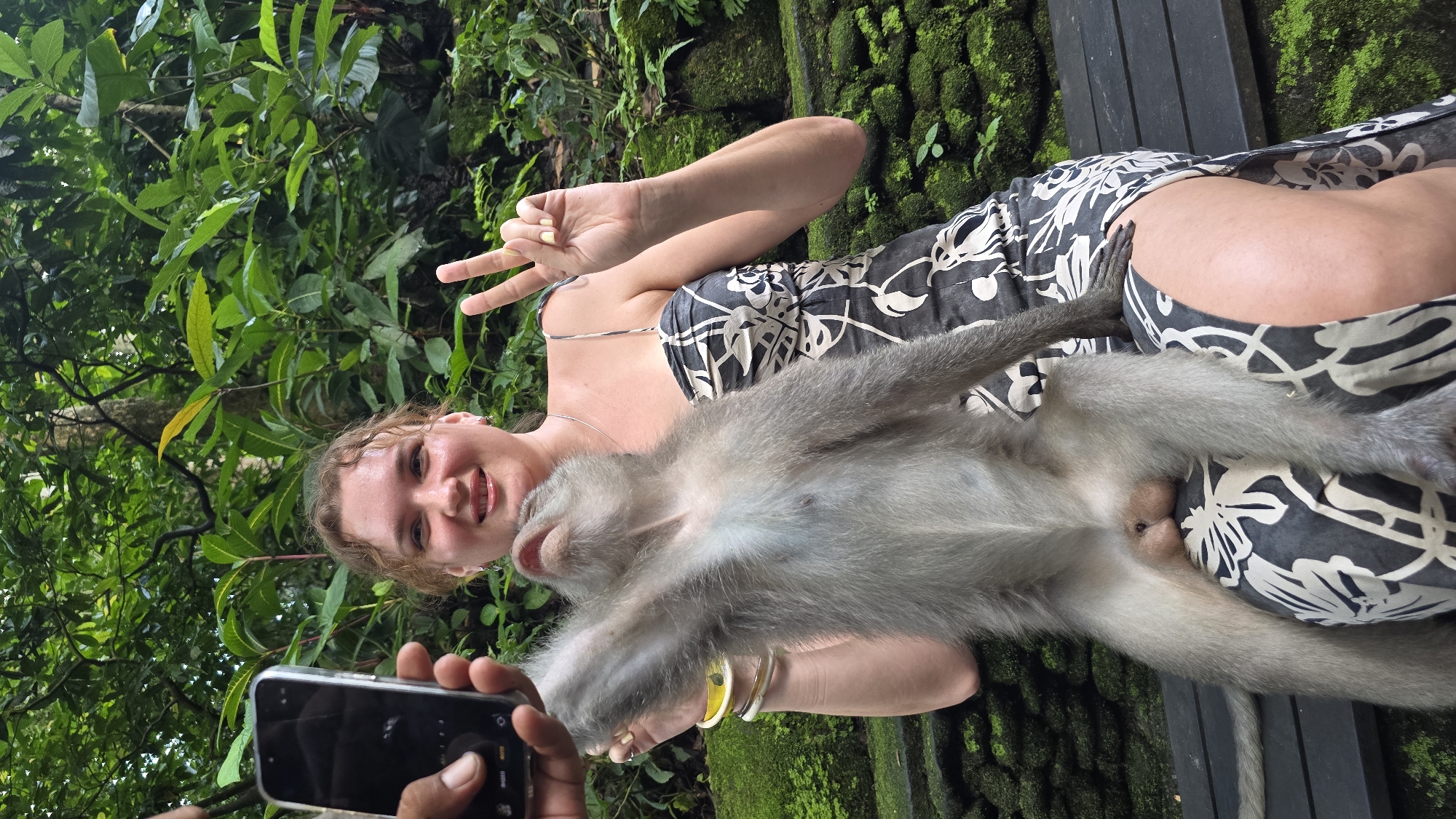 A photograph taken in the Ubud District, Indonesia, depicts a Caucasian woman with curly, light-brown hair wearing a black-and-white floral-patterned sundress. She is smiling at the camera and making a "peace sign" with her right hand. A gray monkey rests on her lap. The monkey's belly is facing the camera. A person in the foreground holds up a smartphone, presumably to take the photo. Lush, green foliage and moss-covered walls form the background. A wooden bench is visible behind the woman.