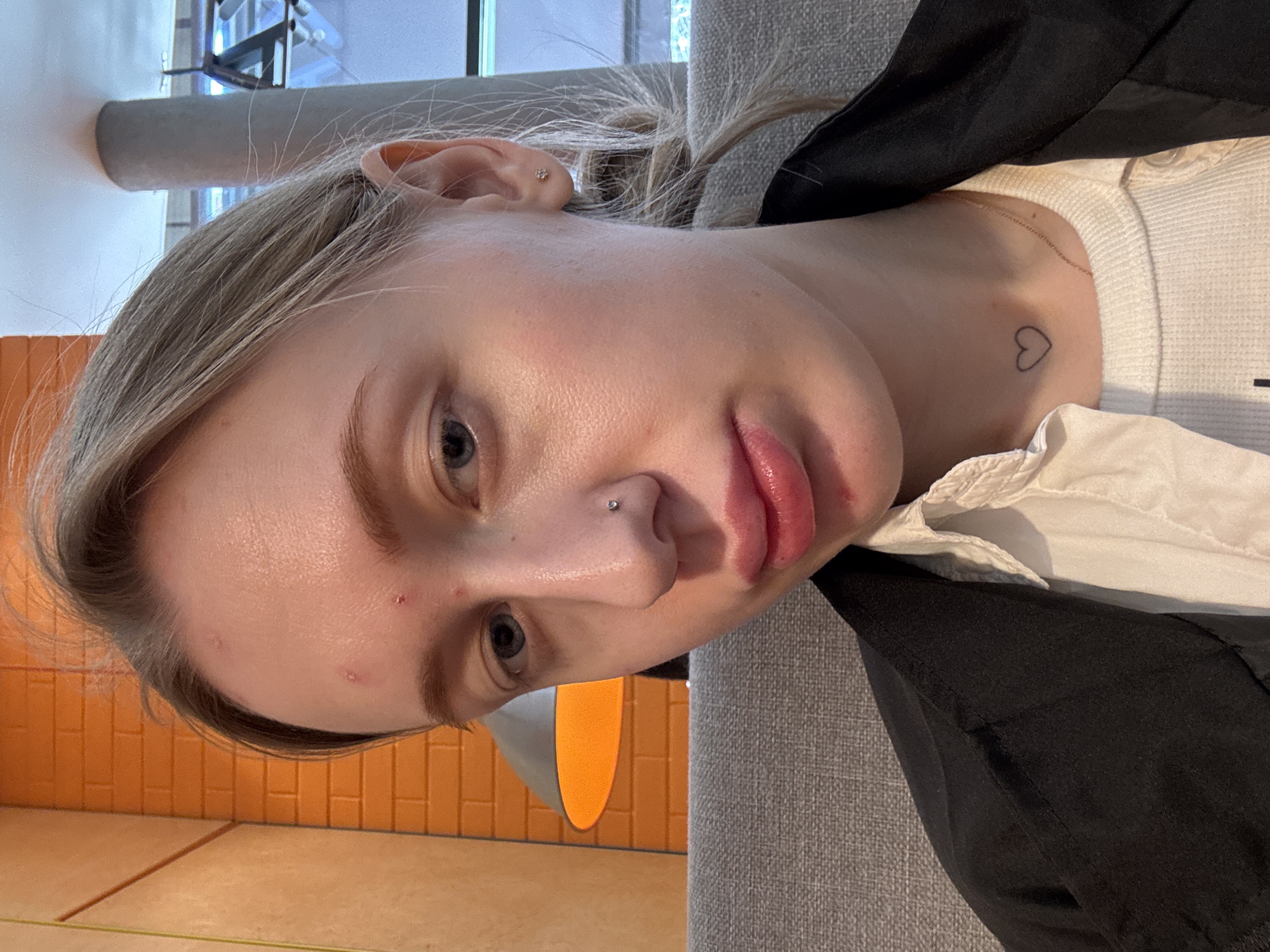 A close-up, rotated image of a fair-skinned individual is presented. The subject's hair is light blonde, and their eyes are blue-grey. Facial features include a nose piercing and blemishes on the forehead. A small heart tattoo is visible on the upper chest. The individual wears a white top, partially covered by a black garment. The background includes an orange tiled wall and a gray upholstered surface. The image does not provide enough information to determine a specific location in Rostov-on-Don, Russia.