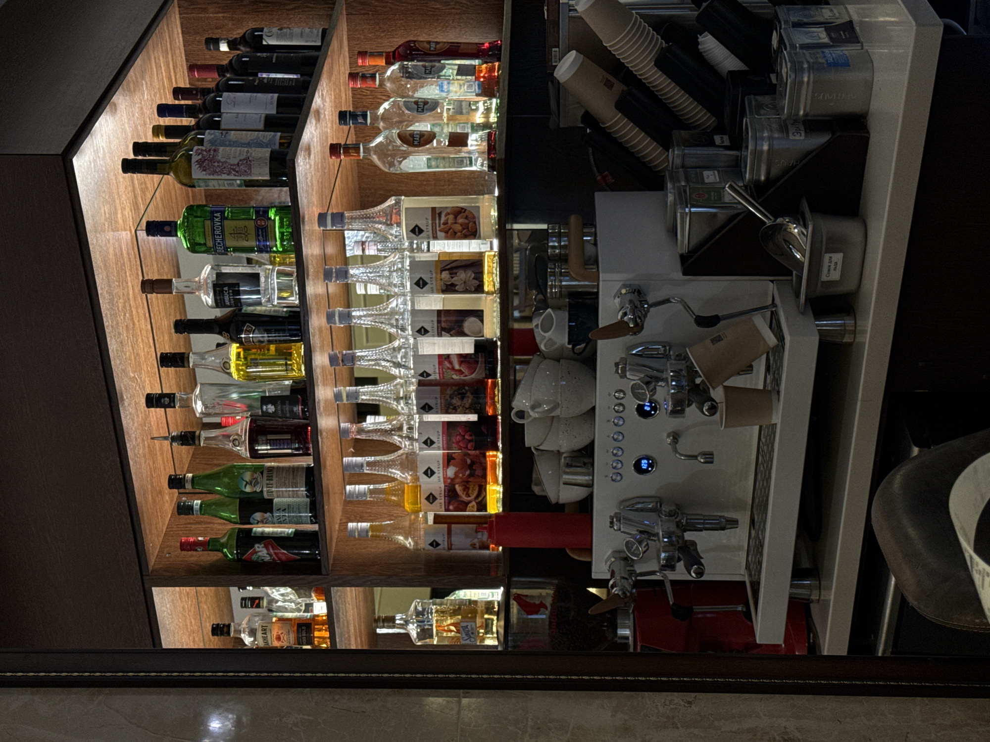 The image depicts a well-stocked bar area, likely in a commercial establishment. On the shelves above, a variety of liquor bottles are neatly arranged. These include clear bottles, some with amber-colored contents, green bottles, and darker bottles that likely contain red wine. Beneath the liquor shelves, a row of syrup bottles is visible, featuring various colors and labels suggesting different flavors such as strawberry and almond.
In the foreground, a professional-grade espresso machine is the central focus. It is a silver and white appliance with various chrome-plated levers and dispensers. To the left of the espresso machine, a coffee grinder is partially visible. A stack of white cups rests on top of the machine. The counter area below the machine is organized with metal containers, some holding what appear to be coffee-related supplies, and a scoop. Disposable coffee cups are neatly stacked in a holder to the right of the machine.
The overall environment appears to be clean and organized, suggesting a functional bar or cafe. There are no individuals present in the image. The setting is indoors, illuminated by artificial lighting that highlights the products on the shelves and the equipment on the counter. The presence of various types of beverages and coffee-making equipment indicates a place that serves drinks. The mention of Urlu-Aspak, Russia, provides a geographical context for this scene.