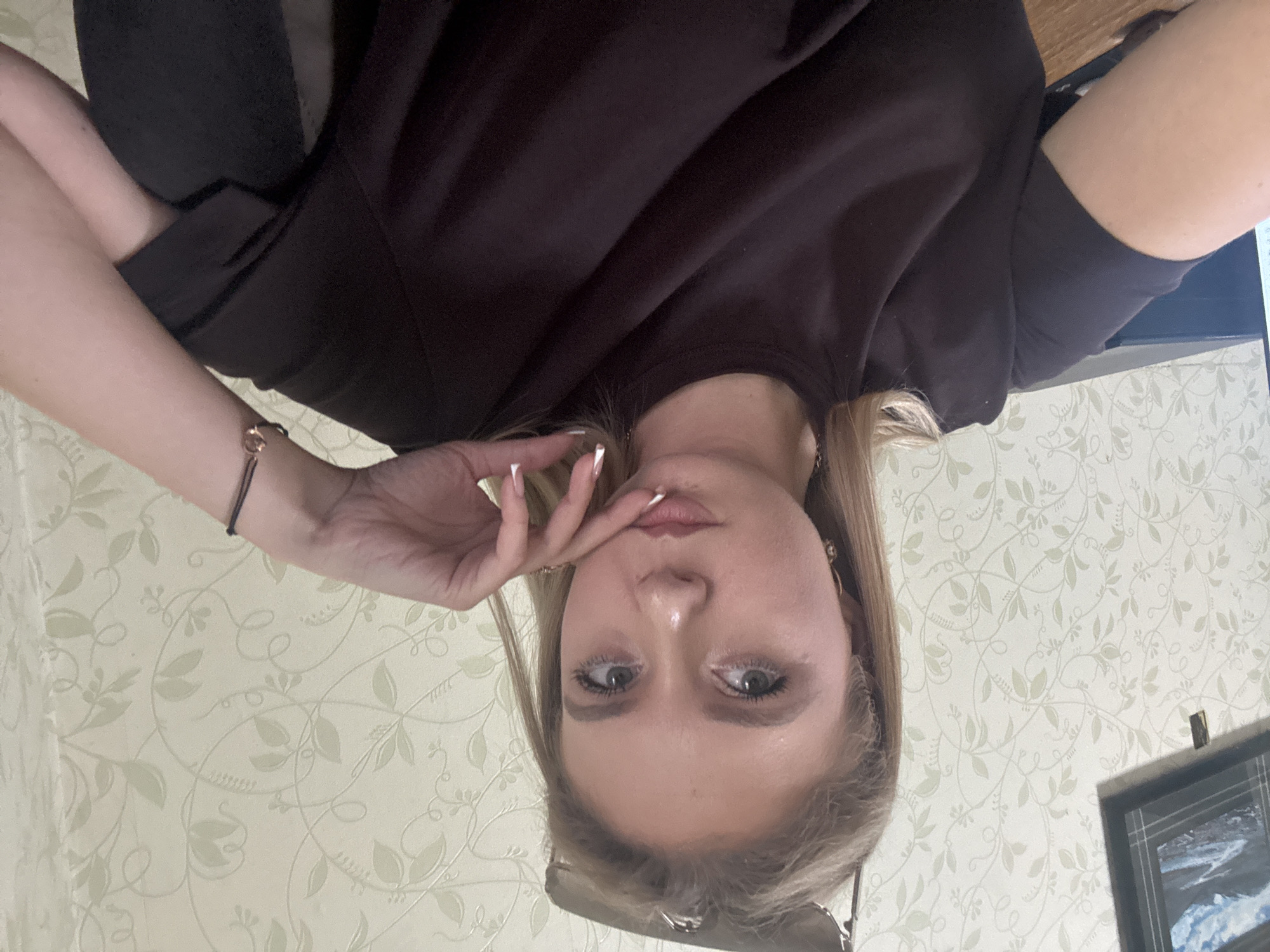 A young woman with blonde hair and green eyes is shown in a close-up, self-portrait style image. She is wearing a dark-colored t-shirt and has her hand resting on her chin, with her index finger extended. Sunglasses are perched on top of her head. The background consists of a wall with a light-colored, floral-patterned wallpaper. A framed picture is visible on the wall to the left of the woman, and a black electronic device is partially visible on a desk in the lower left corner. The lighting suggests an indoor setting.