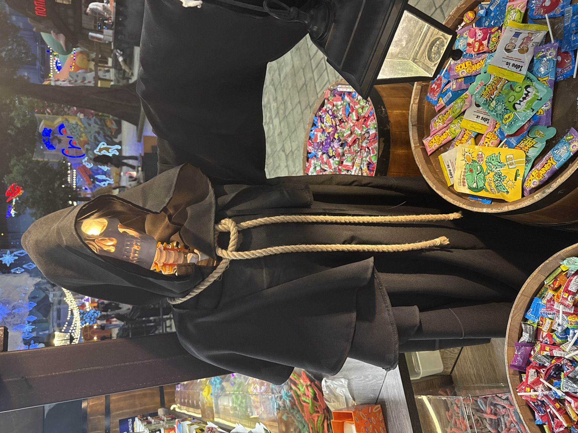 A life-sized Grim Reaper Halloween decoration, complete with a black hooded cloak secured with a rope around its neck, stands adjacent to barrels overflowing with candy, most likely in Moscow, Russia. The city is suggested by the Cyrillic text (ПОП) visible on a lollipop wrapper within the barrels of sweets. Additional barrels and dispensers are seen below the Grim Reaper. Light reflections on the floor and windows suggest it is night.