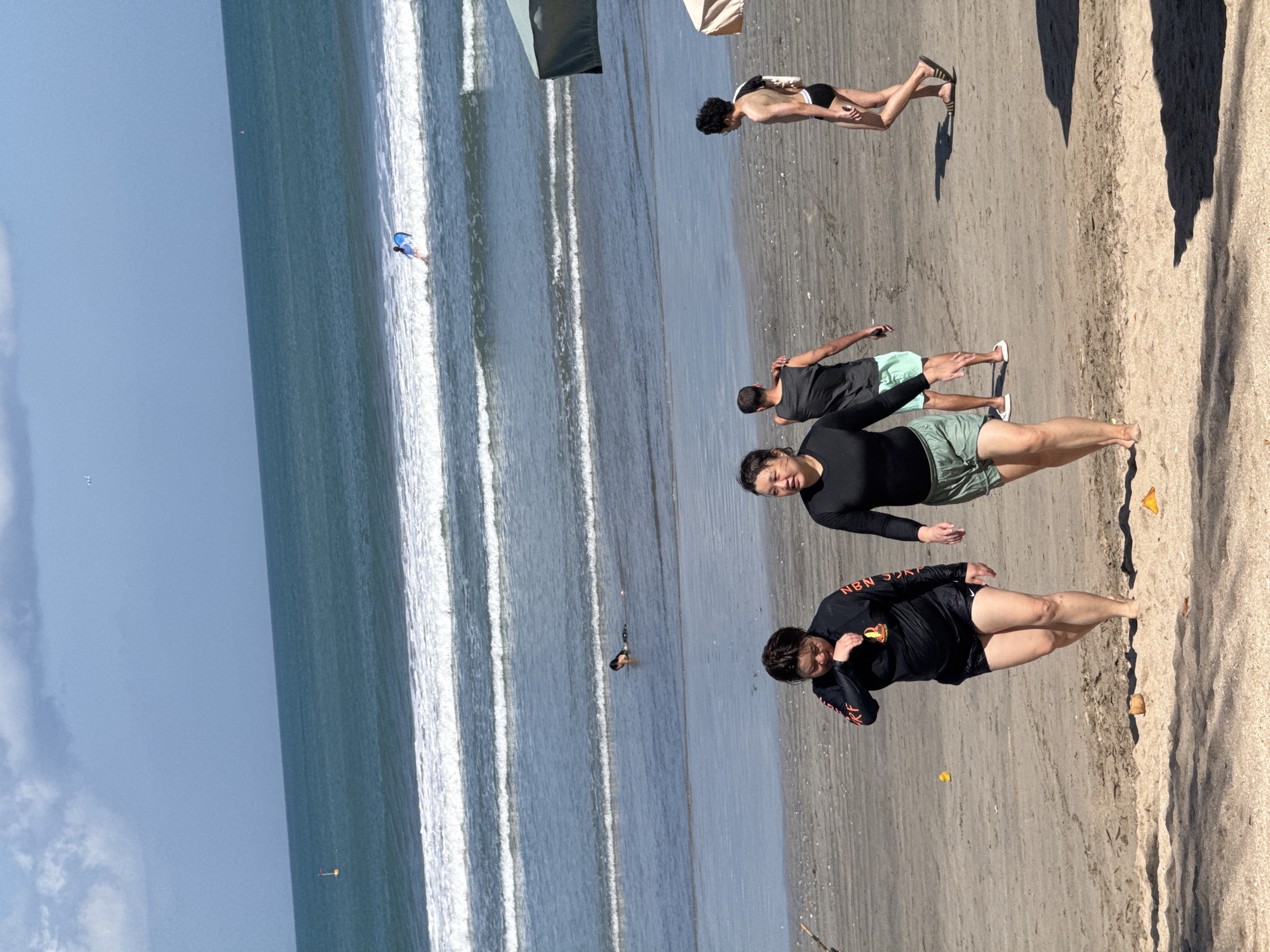 In Kuta, Indonesia, on a sunny day, several individuals are present on the sandy beach and in the ocean. Two women in black long-sleeved tops and shorts are walking on the beach, one touching her face. Behind them, a man in a grey tank top and light green shorts stands, while another man in black swim trunks walks away from the camera. In the ocean, waves are rolling in, and a person is seen bodyboarding in the distance. A few beach umbrellas are visible, casting shadows on the sand.