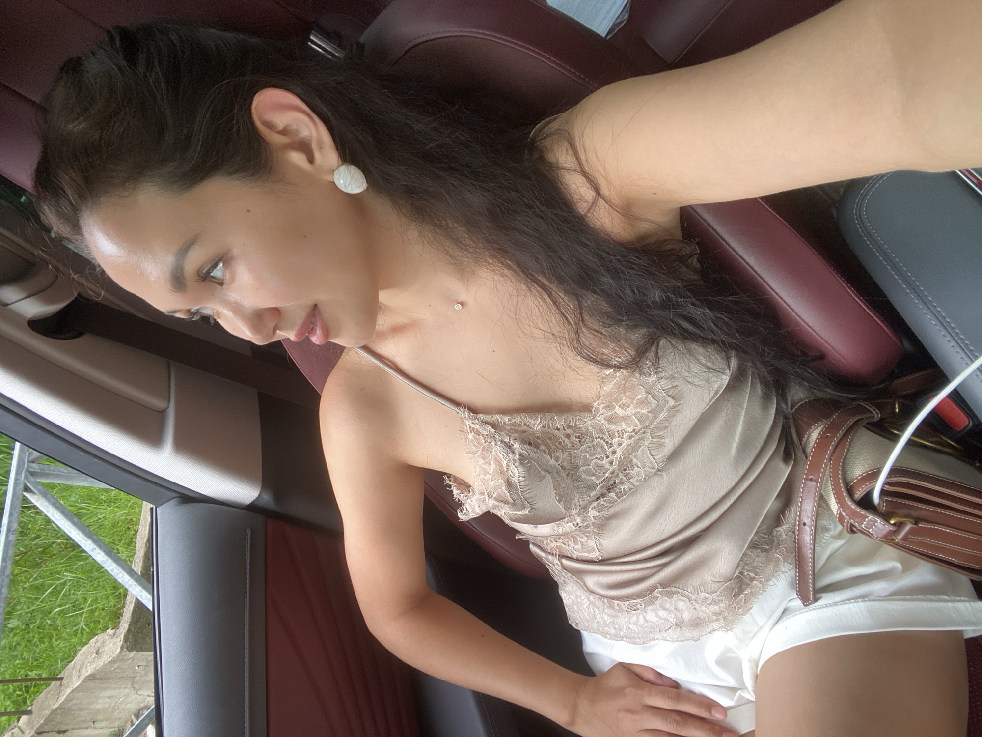 A woman is sitting in the passenger seat of a car. She has dark hair, fair skin, and is wearing a light-colored camisole and white shorts. She is also wearing large, oval-shaped earrings and a small necklace. Her left arm is resting on her lap, and her right arm is extended, suggesting she may be taking a selfie. A brown and tan handbag is visible on her lap. The interior of the car appears to be upholstered in dark red and gray leather. Through the window, a glimpse of green grass and a metal railing is visible, suggesting an outdoor setting. The image is taken from a high angle, looking down at the woman. The location of Vysokoye, Russia is not discernable from the image.