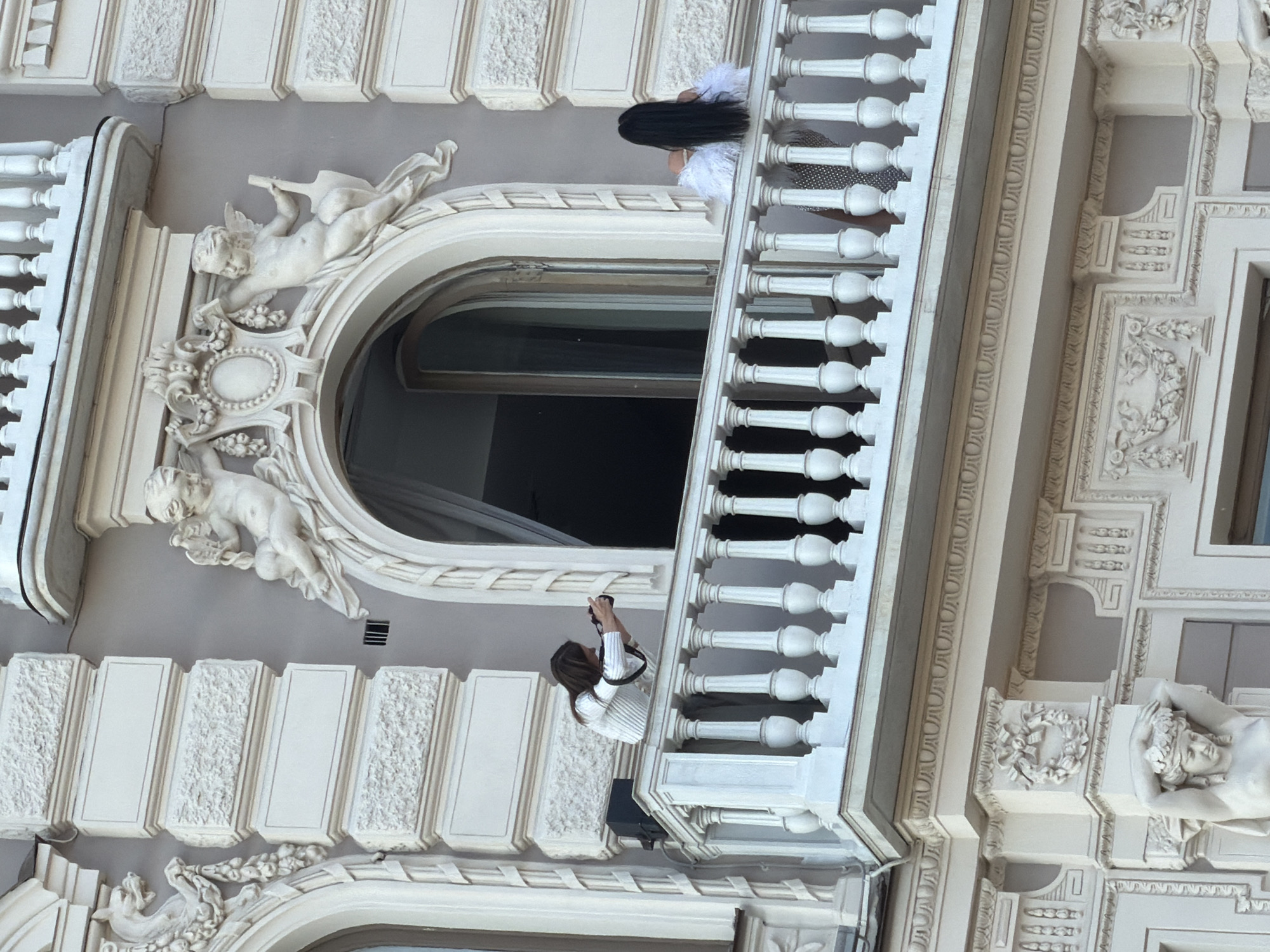 The image shows the façade of a building in Saint Petersburg, Russia, with ornate architectural details, including sculptures of cherubs and decorative moldings. Two people are visible on a balcony: one with long, dark hair and a white feathered top, and another holding a camera, possibly taking pictures. The balcony has a white balustrade with classic design elements. The building's exterior is painted in shades of white and gray.