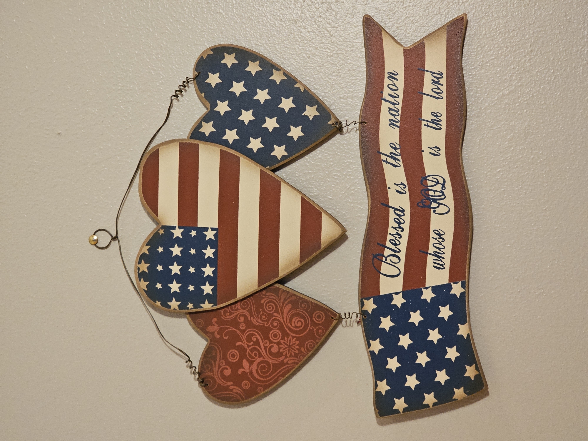 A patriotic decorative wall hanging hangs on a light-colored wall. The hanging features three heart-shaped ornaments and a banner, all in the style of the American flag with red and white stripes and blue fields with white stars. The leftmost heart has a red scroll pattern. The banner displays the text "Blessed is the nation whose God is the lord" in blue cursive lettering. The objects are suspended by wire and attached to a single hook on the wall. The environment is an indoor setting. No people are present.