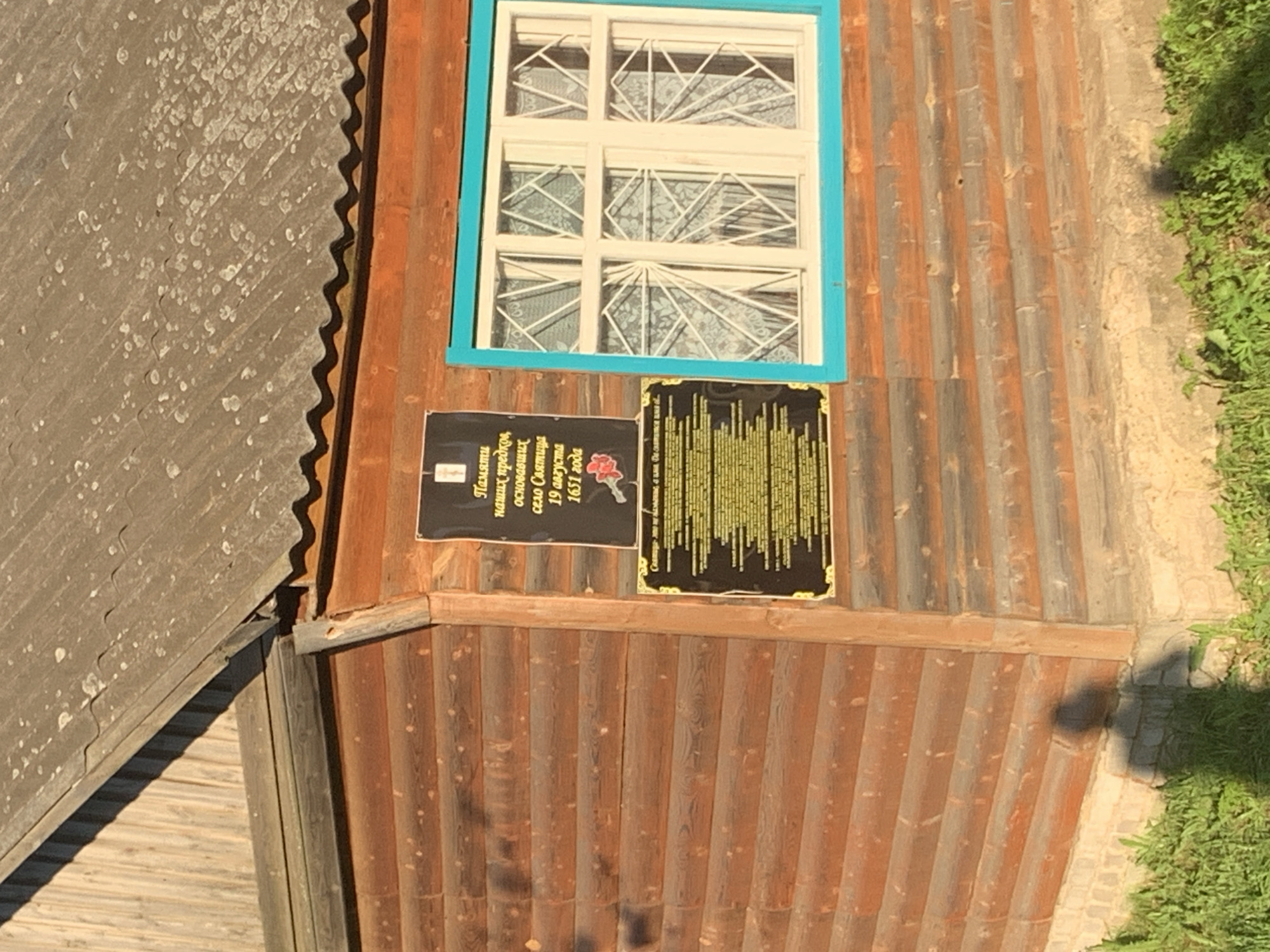 The image shows a wooden building with two plaques attached to its exterior wall. The building appears to be made of horizontal logs. A window with a blue frame is visible to the right of the plaques. The plaques are black with gold lettering and designs. The top plaque has text that appears to be in Russian, with a red rose image at the bottom. The bottom plaque also contains extensive Russian text. The scene is outdoors, with green grass and sunlight suggesting a daytime setting. A shadow of a person is visible on the ground near the base of the building. The plaques are indicative of a historical marker or dedication, possibly related to the founding or significant events in the village of Святица, Russia.