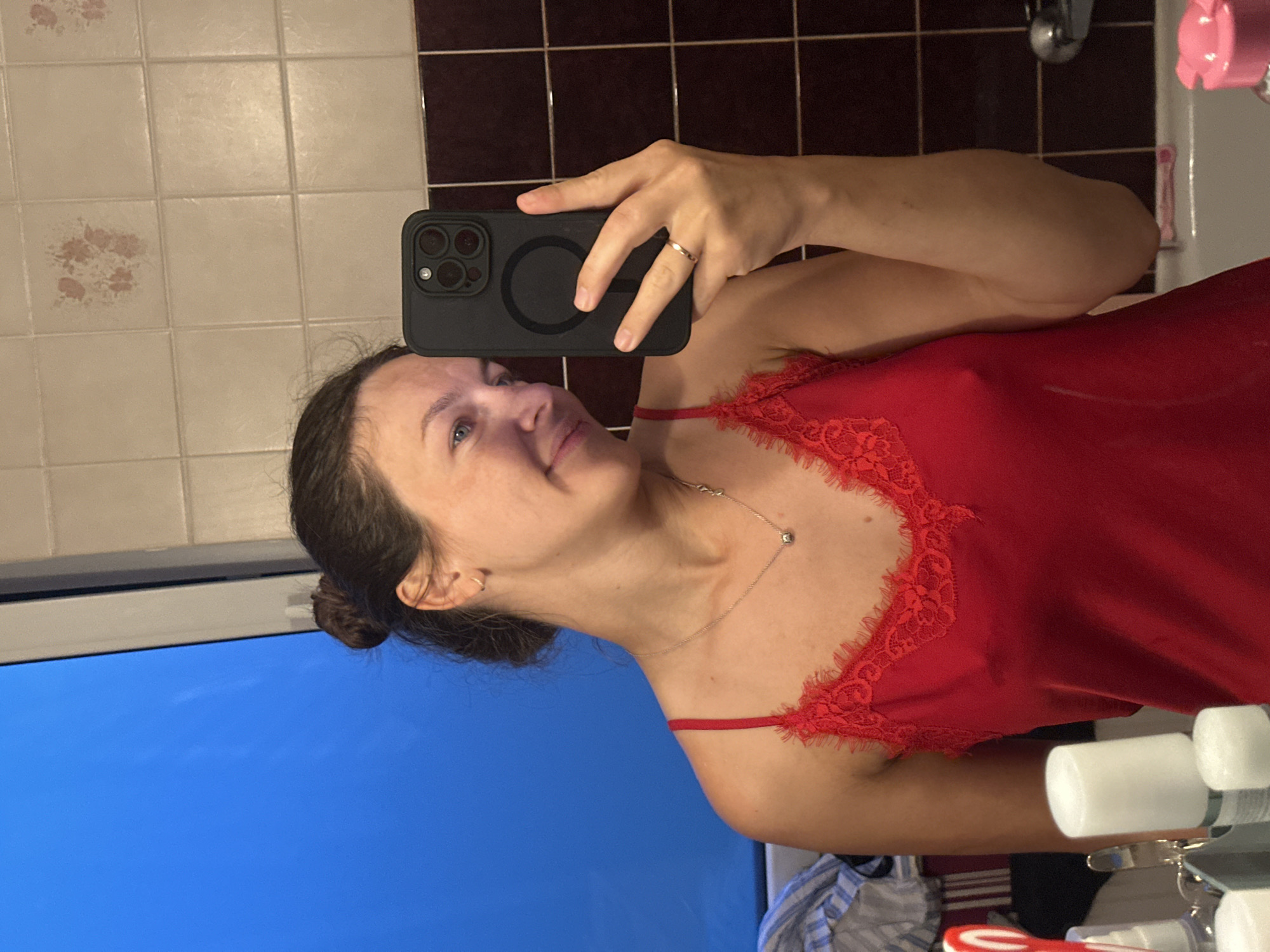 A woman is depicted taking a mirror selfie in what appears to be a bathroom. She has dark hair pulled back into a bun, light eyes, and a slight, pleasant smile. She is wearing a red camisole or nightgown with delicate lace trim along the V-neckline and thin shoulder straps. A thin silver necklace with a small charm hangs around her neck, and she wears a simple ring on her right ring finger, which is also visible holding the smartphone. Small hoop earrings are visible in her ears.
The woman holds a black smartphone, featuring a distinctive multi-lens camera module and a circular magnetic attachment (likely MagSafe) on its case, up to capture her reflection. The setting is a tiled bathroom. The reflected walls show a pattern of square tiles; the upper section features lighter, possibly beige or off-white tiles, some with faint reddish decorative patterns, while the lower section has darker, possibly maroon or brown tiles.
Parts of a white sink basin, a silver faucet, and various toiletries in white bottles are visible on a counter to the right. A pink item, possibly a cup, is also near the faucet. To the left, a large blue reflection, possibly from a screen or window, fills the lower portion of the image. The lighting suggests an indoor setting, likely with artificial light. No legible text is visible in the image.