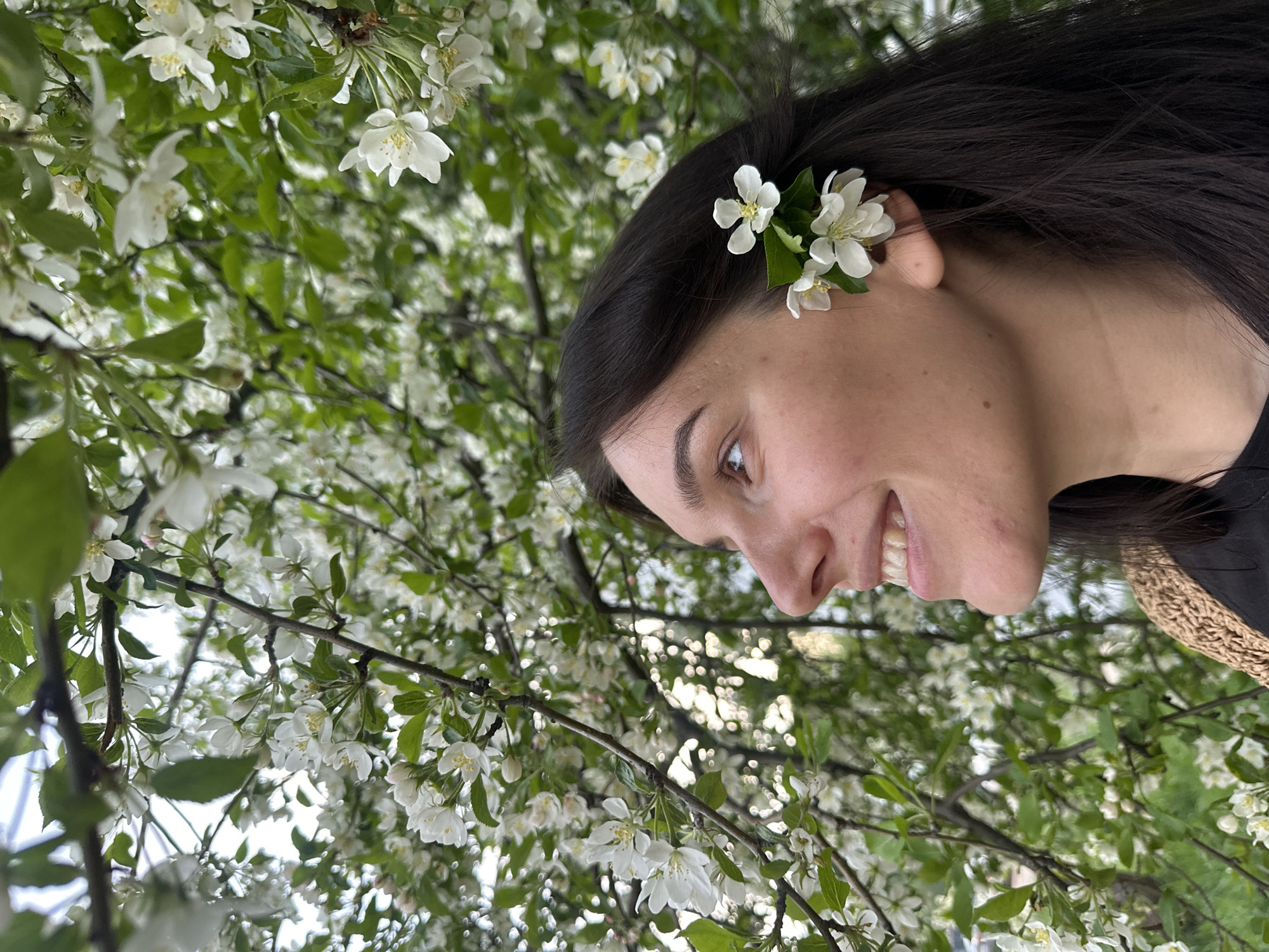 A woman with dark hair and fair skin is pictured in profile, smiling, with white flowers tucked behind her ear. The background is filled with a tree abundant with white blossoms and green leaves. The location is potentially Chaikovsky, Russia, but there is no confirmation. No other individuals are present.