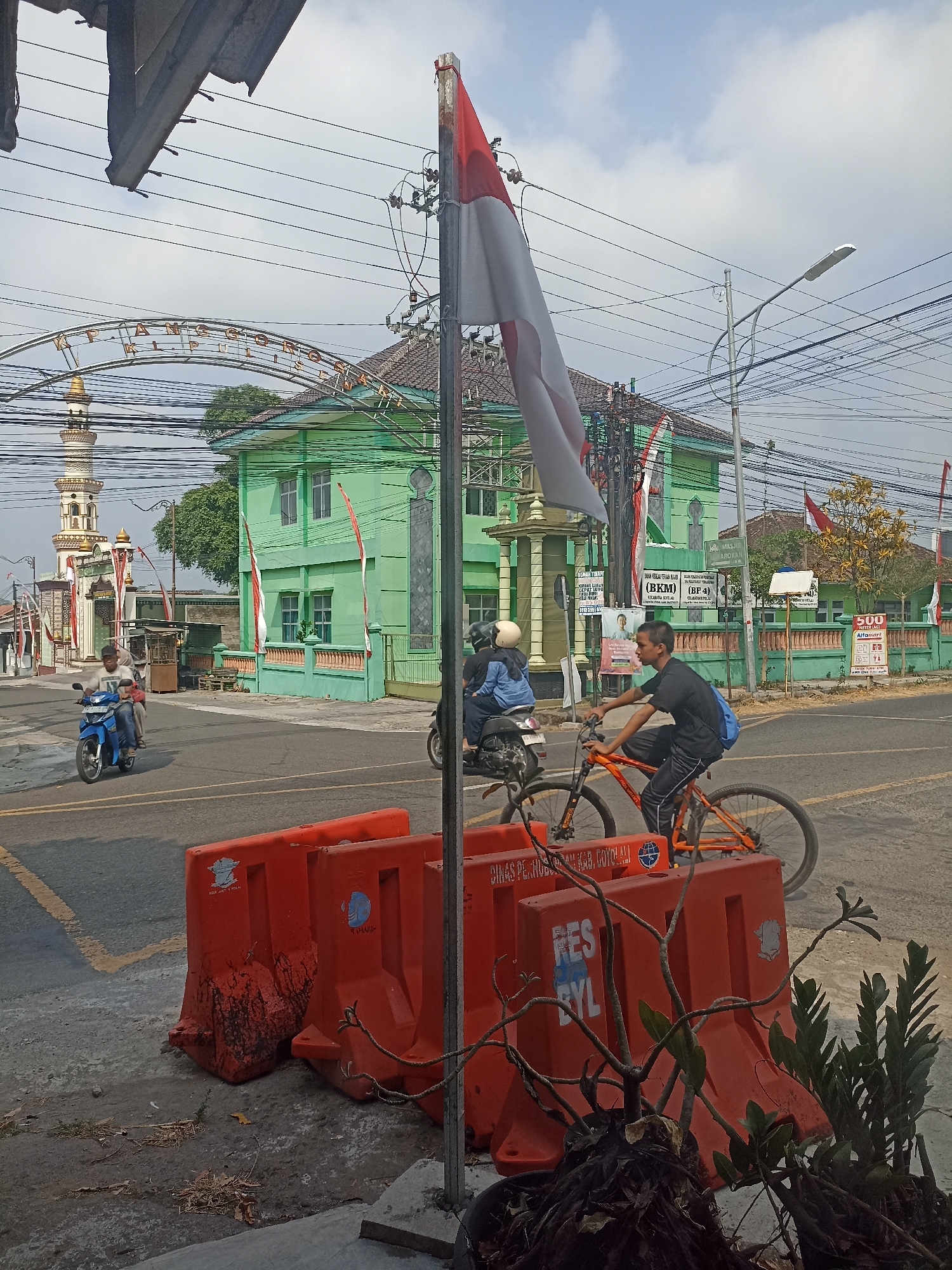 A street scene in Pulisen, Indonesia, shows a bright green building with a mosque-like minaret on the left. A prominent Indonesian flag, red and white, waves from a pole in the foreground. Several power lines crisscross the sky above. In the street, a man rides an orange bicycle, and another person is on the back of a blue motorcycle. Orange plastic barriers are placed on the side of the road, some with debris on them. The weather appears to be daytime with a cloudy sky.