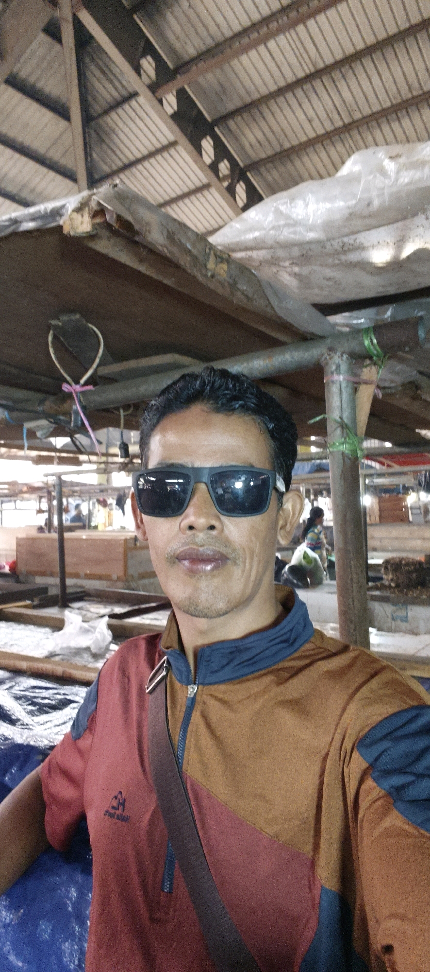 A man with dark hair and a mustache, wearing sunglasses and a color-blocked jacket with red, brown, and navy blue sections, stands in what appears to be an outdoor market or covered workshop. The man is taking a selfie, with his face taking up a significant portion of the foreground. He has a dark strap across his chest, part of a bag. In the background, there are wooden structures, tarps, and other people visible, suggesting a bustling commercial environment. The overhead structure is made of metal and wood, with corrugated roofing. The scene is consistent with the general appearance of markets in Jambi, Indonesia, known for their active trade and open-air or covered market setups.