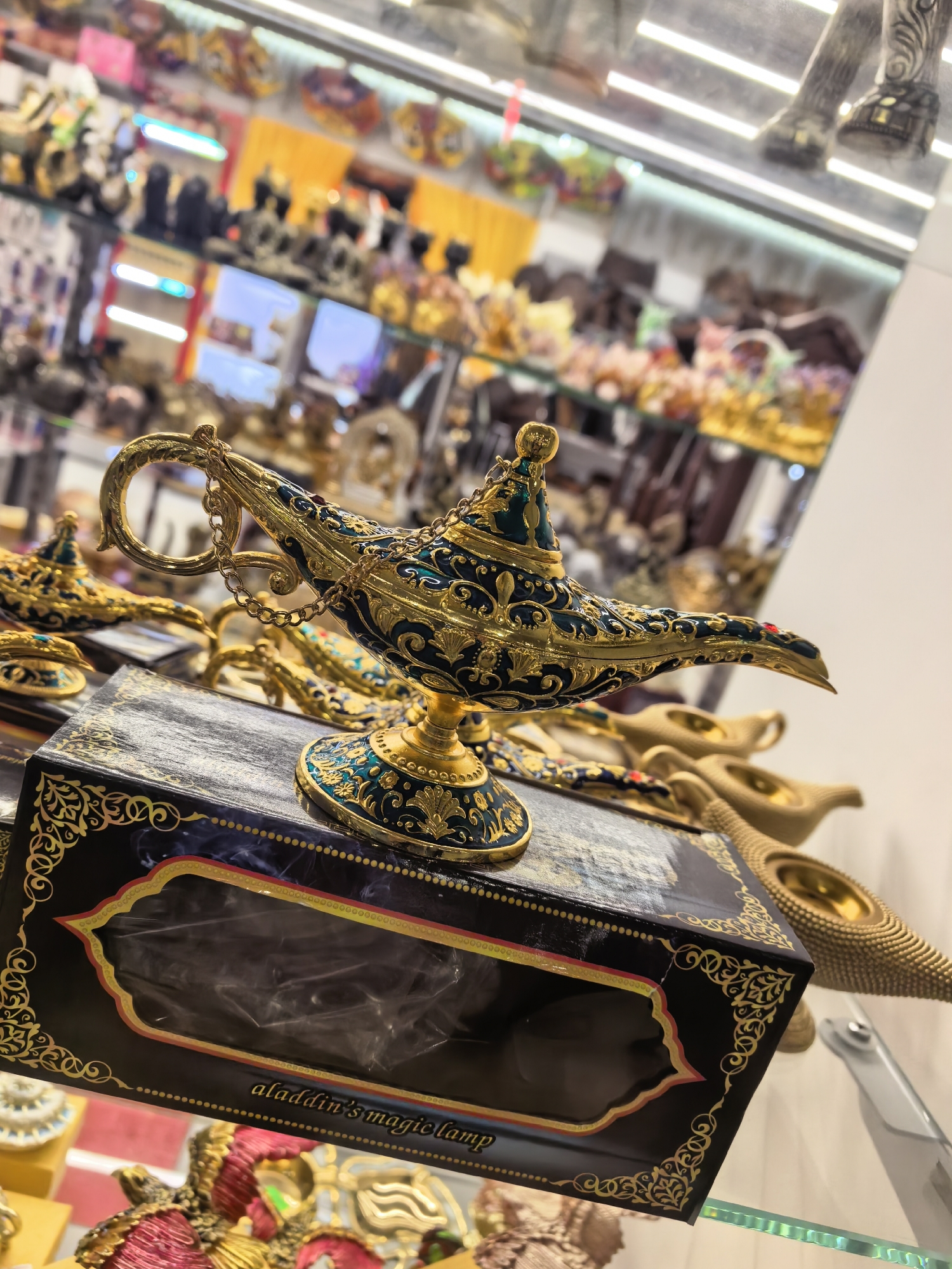 An eye-level, medium shot features a blue and gold "Aladdin's Magic Lamp" on display in a shop. The ornate lamp is centrally located, and sits on a dark box with an ornate gold design. The box has a cutout window and the phrase "aladdin's magic lamp" printed on the side. Other similar lamps are visible in the background, along with various other items on shelves that extend into the distance. The objects are within a glass display case. Based on available information, it is likely that this image was taken in a shop in Moscow, Russia.