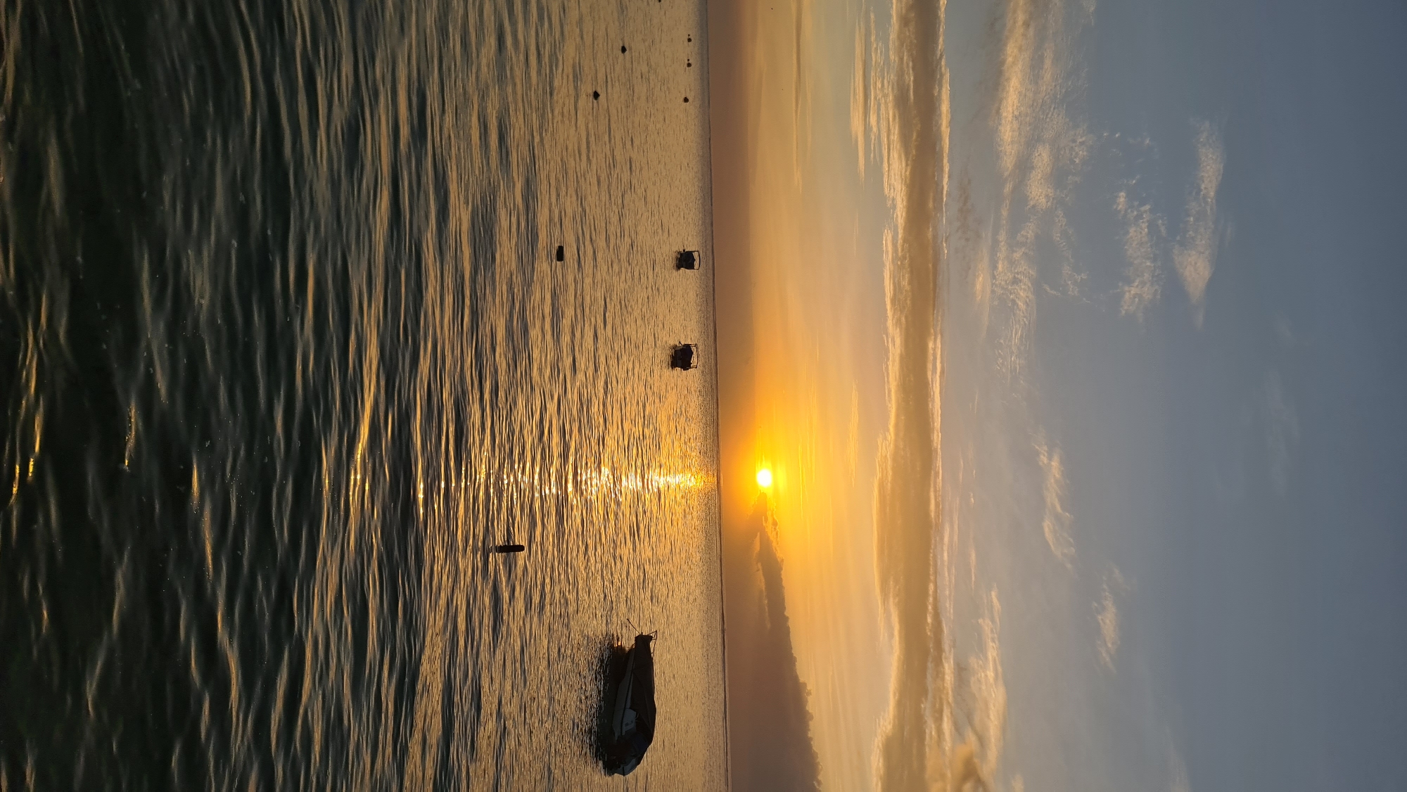 The image depicts a serene sunset over the ocean, likely near the coast of Denpasar, Indonesia. The sun, a bright orb, is low on the horizon, casting a golden glow across the sky and reflecting a shimmering path on the water's surface. The sky is a gradient of pale blue at the top, transitioning to hues of orange and yellow closer to the sun, with scattered wispy clouds tinged with the warm sunset light. The ocean is calm, with gentle ripples disturbing the surface. Several small boats are visible, some appearing to be moored in the distance. On the right side of the frame, a covered boat is partially submerged or very low in the water. A dark, slender object, possibly a buoy or marker, protrudes from the water in the foreground. The overall atmosphere is tranquil and picturesque, characteristic of a tropical sunset.