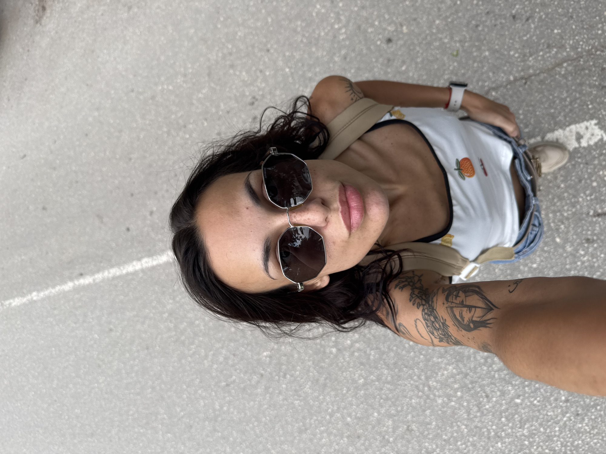 An individual, identified as a woman, is captured in a high-angle selfie on an asphalt surface with a faded white painted line. She has dark hair and wears silver-framed, octagonal Polaroid sunglasses, which obscure her eyes. Her lips are slightly pursed. She is dressed in a white tank top with black trim and an embroidered orange fruit design, light blue denim shorts, and light-colored shoes. A beige backpack strap is visible on her right shoulder. Tattoos are present on both arms: a design featuring two faces within a heart shape on her left arm, and a radial sun-like motif on her right arm. A white smartwatch is worn on her left wrist. The outdoor setting is in the city of Pleven, Bulgaria.