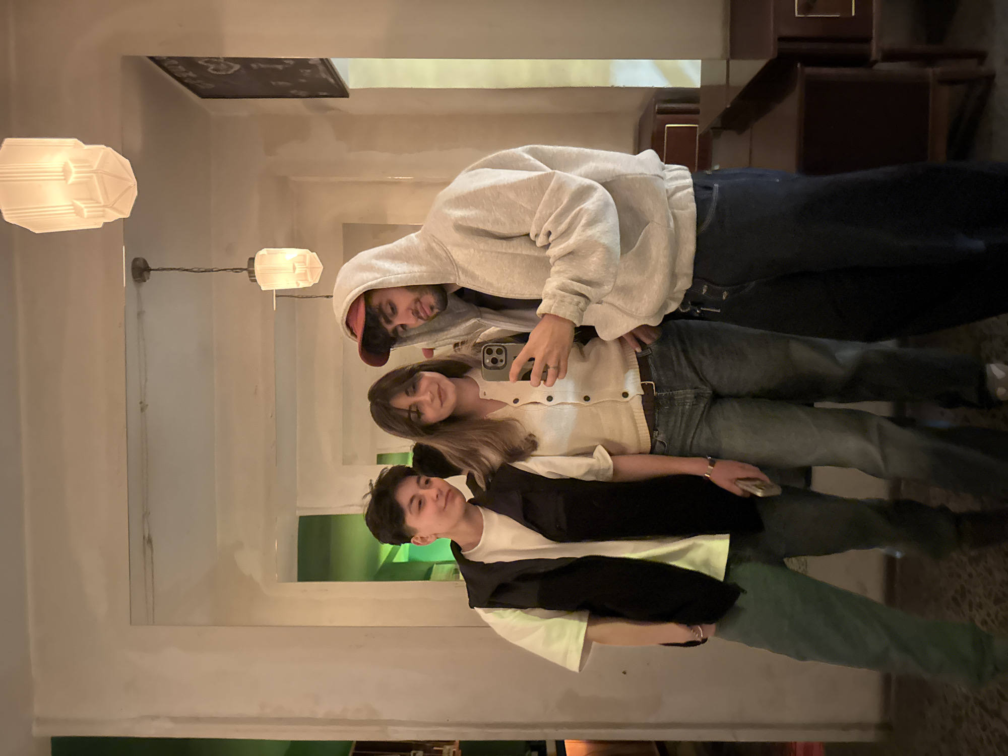 Three people are posing for a selfie. The person in the back is wearing a grey hoodie and red baseball cap, holding a smartphone. The middle person has long brown hair and is wearing a white cardigan. The person in the front is wearing a white t-shirt with a black vest. The location appears to be indoors, potentially in a cafe or bar. Hanging lights can be seen on the left side of the image. The location may be Yerevan, Armenia, although there is no specific information that explicitly confirms the location.