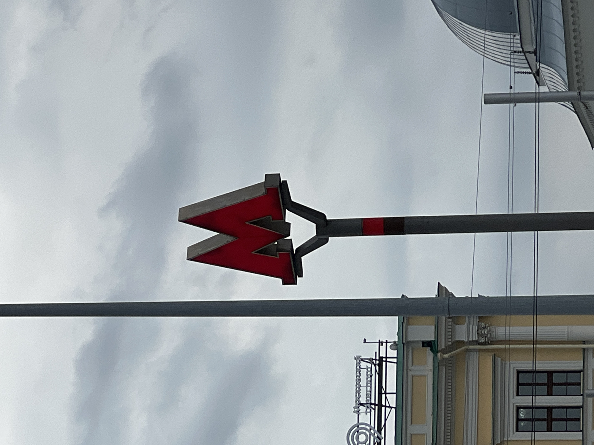 The image shows a red "M" sign for the Moscow Metro, a tall gray pole, and part of a yellow building with classical architectural details. There is a silver dome on a building in the background to the right. The sky is overcast with gray clouds. A sign with the text "Мосгортранс" is visible on the yellow building. The overall scene suggests an urban environment in Moscow, Russia, likely near a metro station.