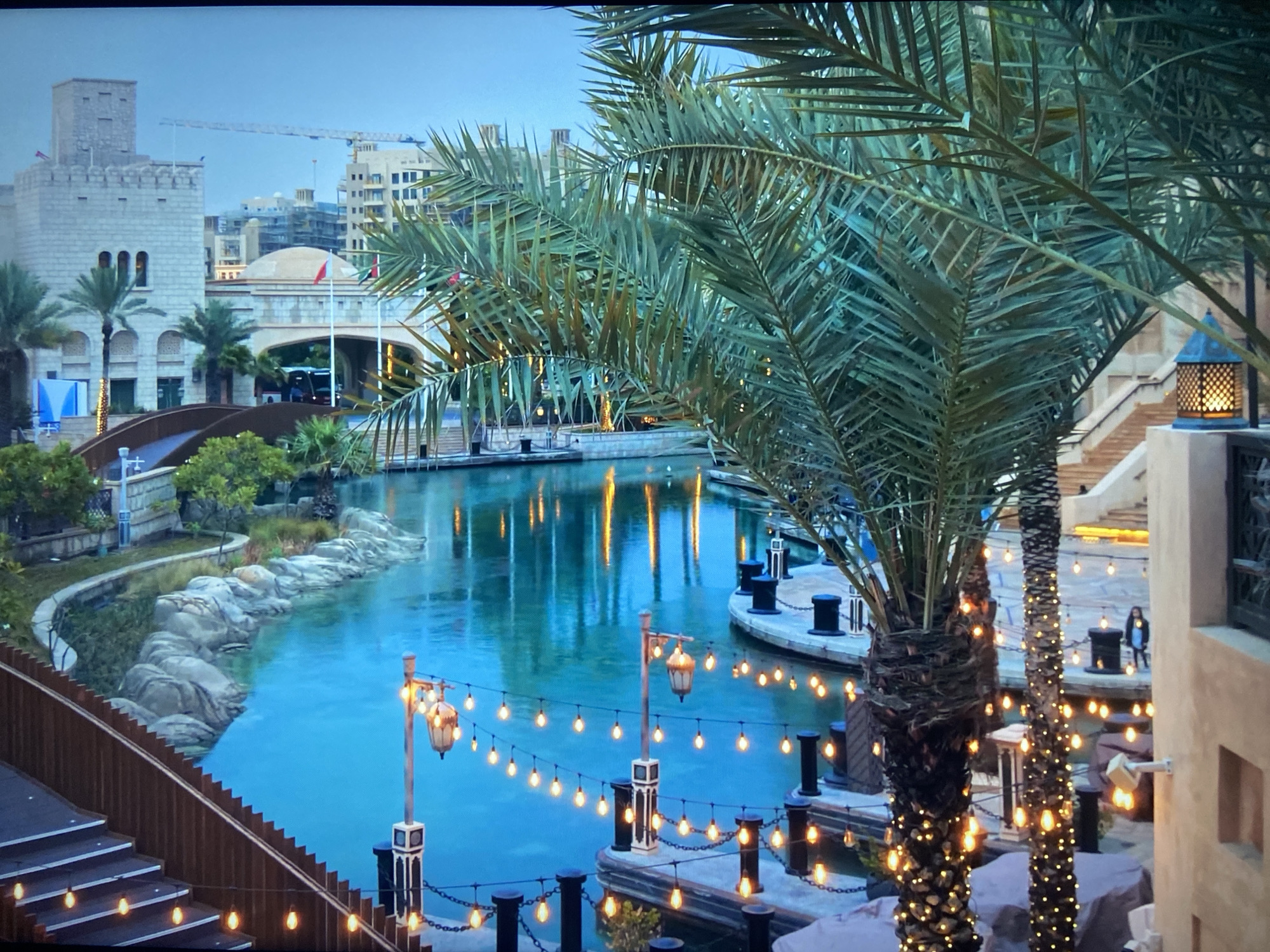 The image shows a canal scene in the city of Unknown, Unknown, featuring architecture reminiscent of a Middle Eastern style. Buildings with arches, crenellations, and domes can be seen. A canal runs through the scene, reflecting the light from street lamps and lights strung along the banks. Two palm trees, decorated with lights, stand in the foreground. Further back, a bridge is visible. The canal is lined with stone features and docks with black bollards. The overall atmosphere is serene, with a warm glow from the lights.