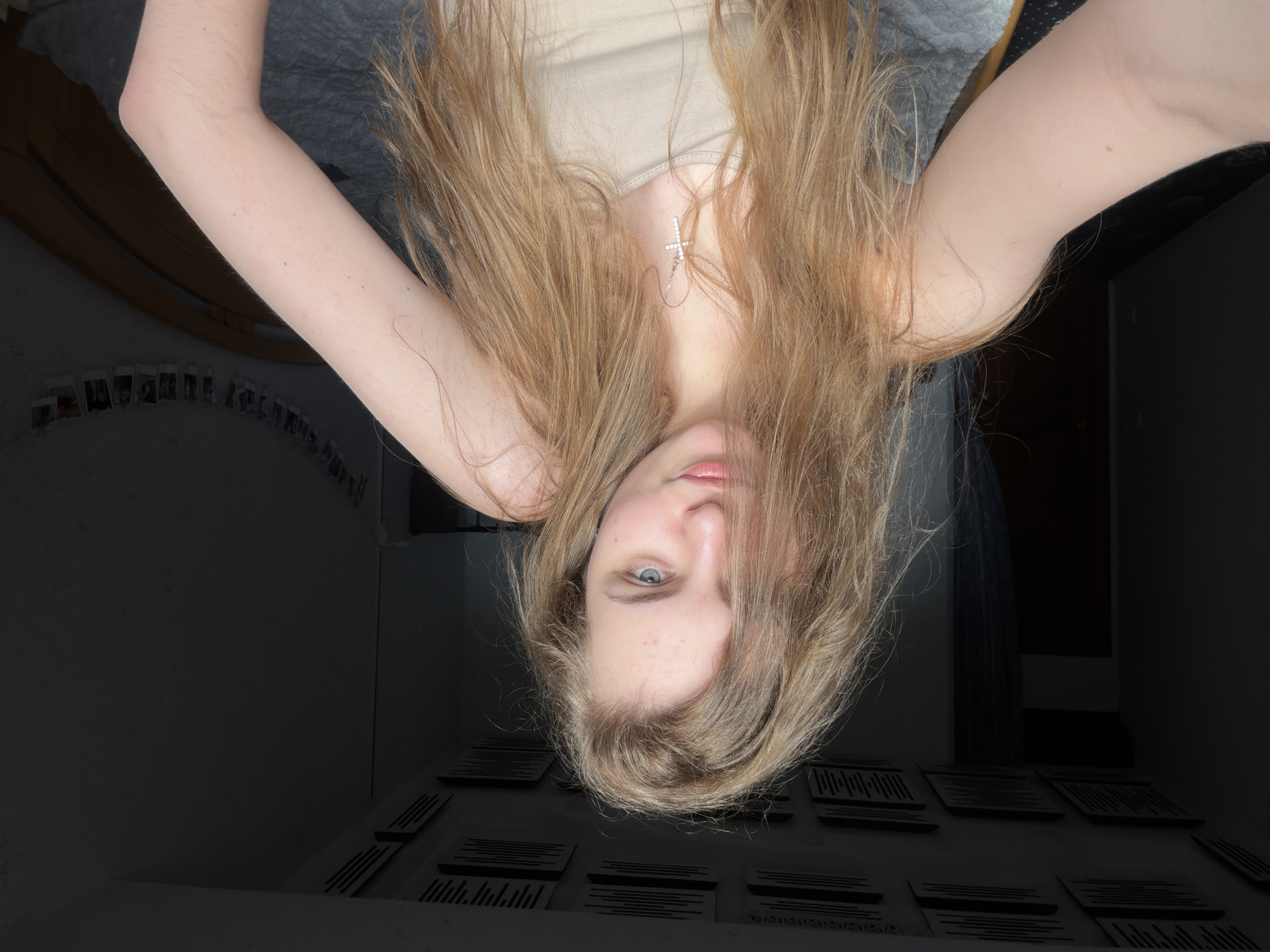 A young woman with long, blonde, wavy hair is pictured in a selfie, looking directly at the camera. She is wearing a light-colored tank top and a silver cross necklace. Her right arm is bent and visible in the foreground. The background shows a room with a dark wall, a bed with gray bedding, and a string of photos hanging on the wall. The ceiling has dark acoustic panels. The image does not contain any discernible landmarks or features that would indicate a specific location within the city of Dnipro, Ukraine.