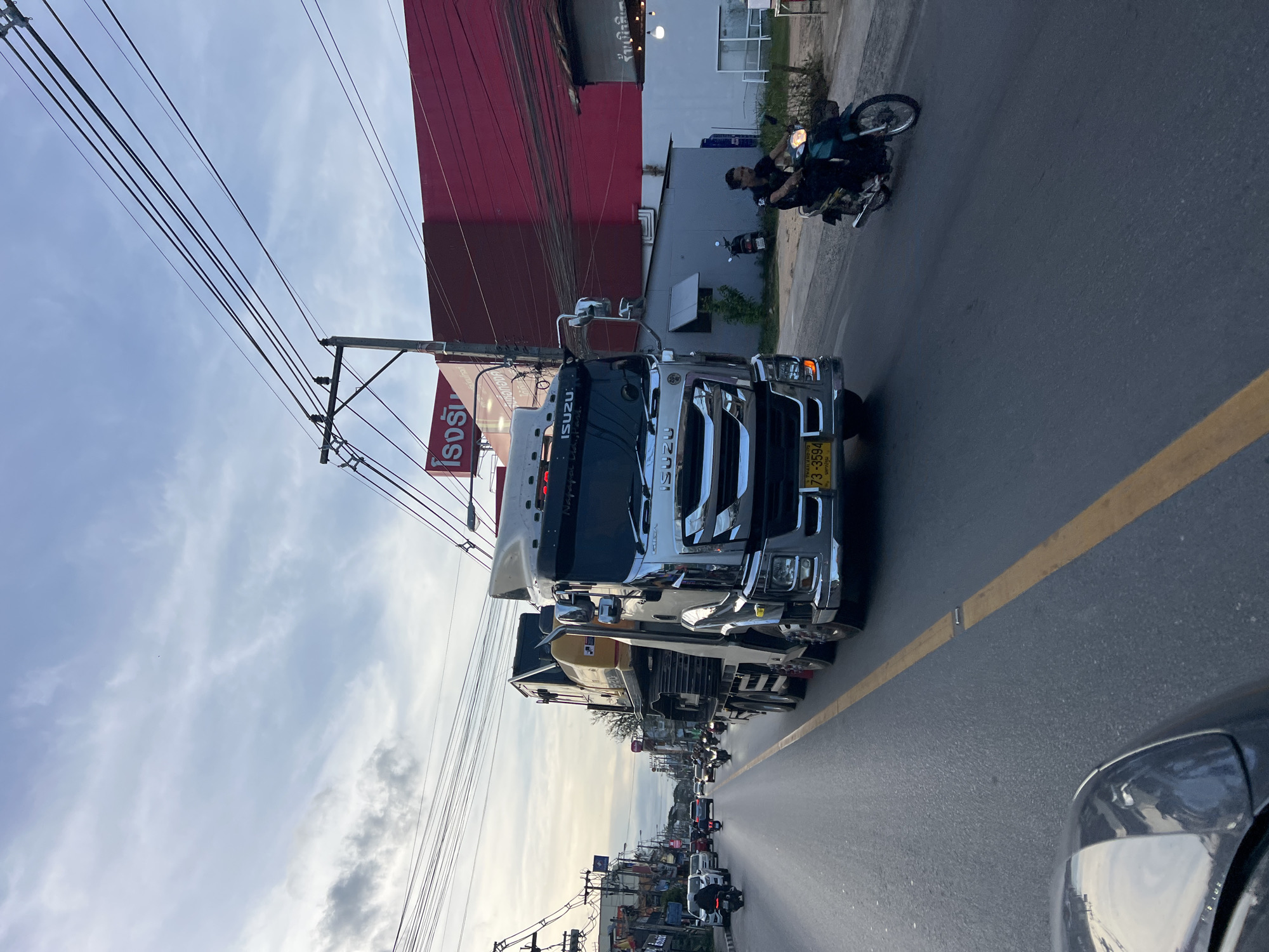 The image captures a street scene in Baan Bo Phut, Thailand. A large, chrome Isuzu truck dominates the foreground, positioned on the right side of the road near a yellow line. A motorcycle with two riders is visible slightly ahead of the truck on the same side of the road. Further down the road, there is a line of vehicles, including more motorcycles and cars. Buildings line the street, with a red-sided building and a sign with Thai script visible near the truck. Overhead, a network of electrical wires stretches across the sky. The sky is partly cloudy. The bumper of a dark colored car is visible in the bottom right corner.