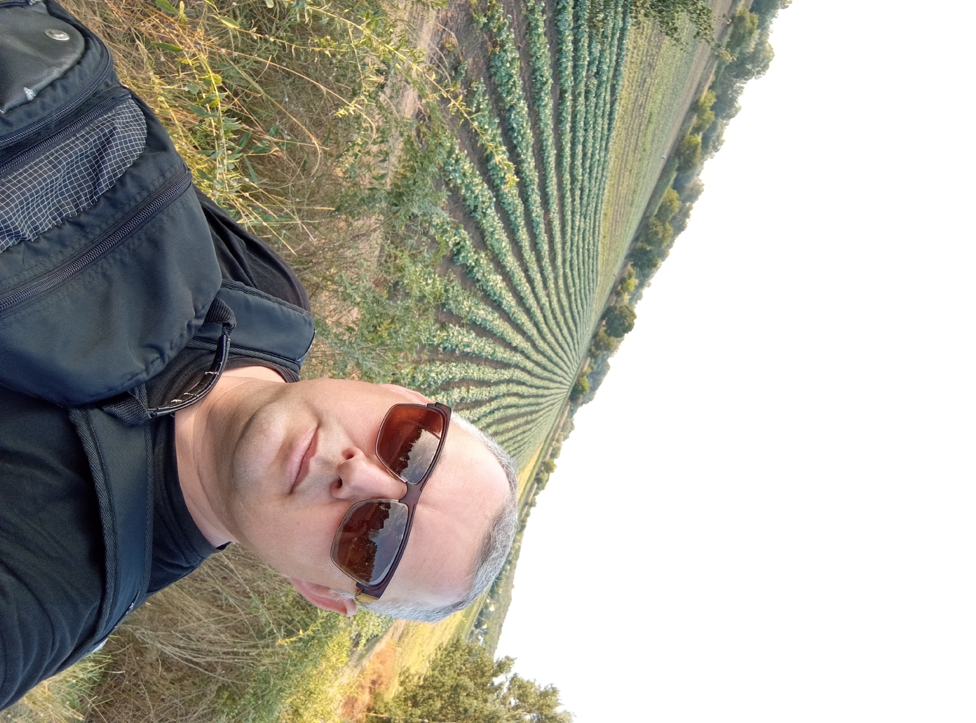 A man wearing sunglasses and a black t-shirt is taking a selfie in front of a field of crops. He is wearing a black backpack. The field is on a hillside, with rows of green plants stretching into the distance. Trees are visible on the surrounding hills. The sky is white and overcast. The location is Mereneshty, Moldova.