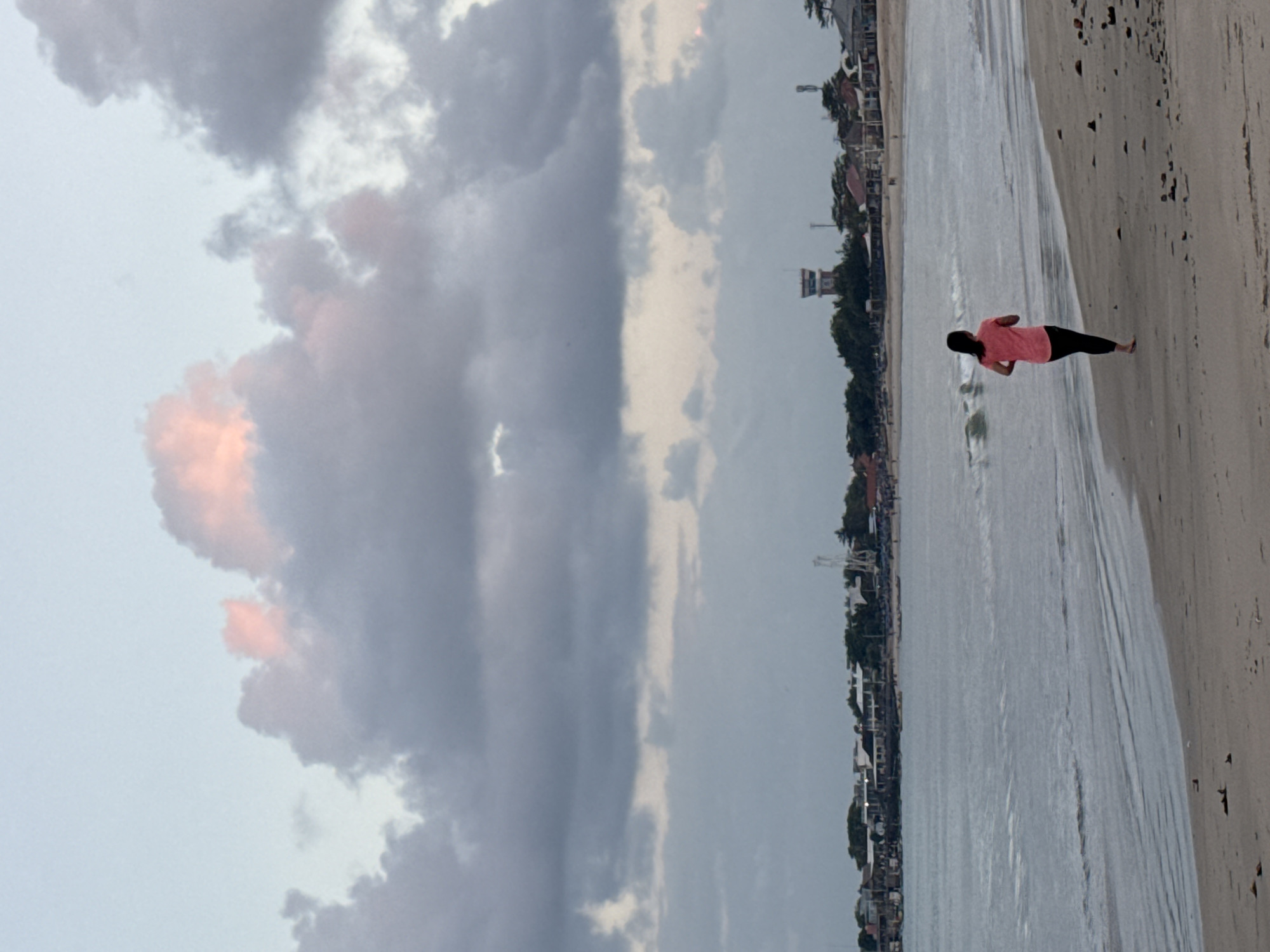 A lone woman in a pink t-shirt and black leggings jogs along a sandy beach. The tide is out, leaving wet sand near the waterline. Gentle waves lap the shore. In the background, a line of buildings, trees, and a control tower suggest a coastal town, likely Kuta, Indonesia. The sky is filled with dramatic clouds, some tinged with pink from the setting or rising sun.